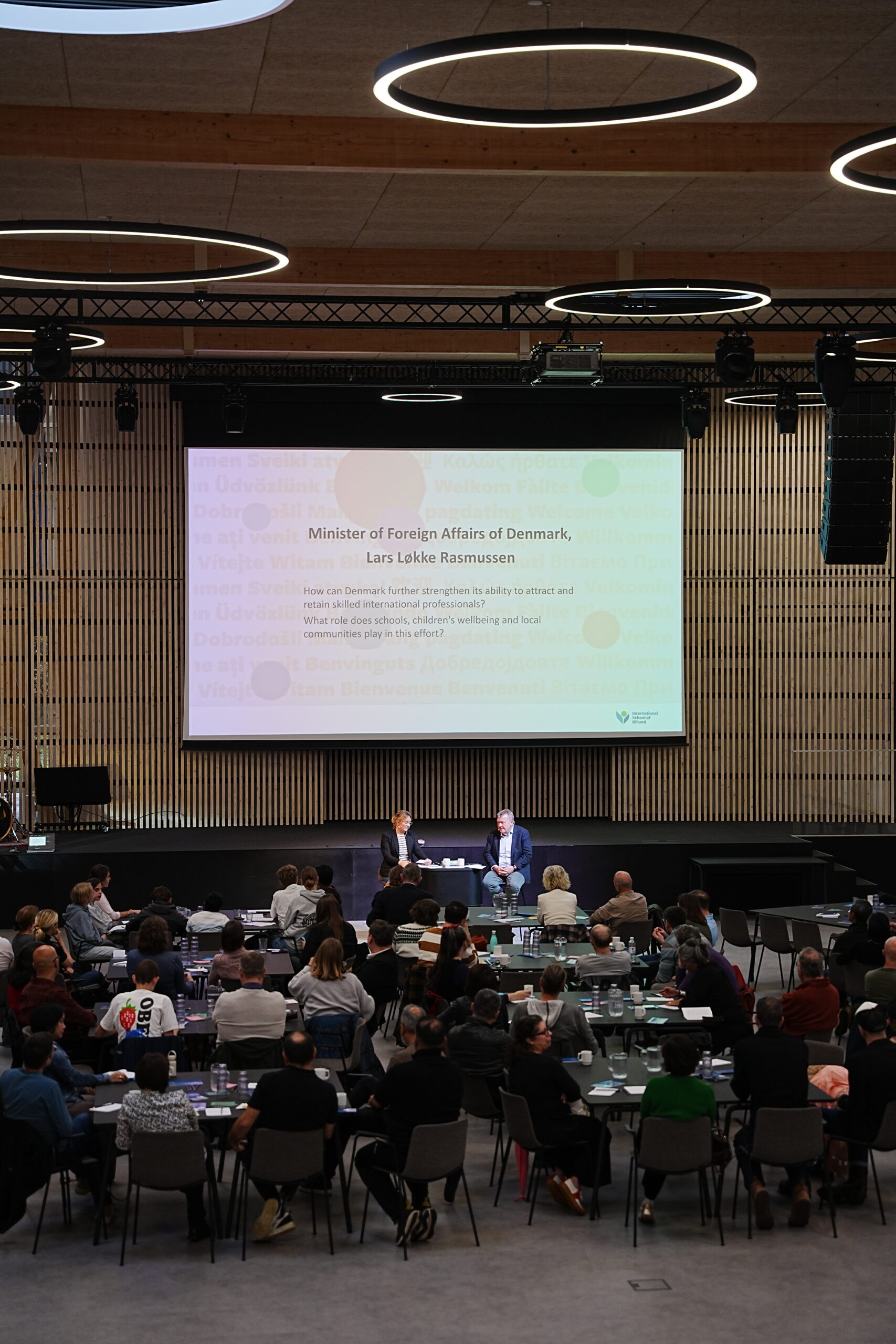 2 men sitting in front of an audience with a projector screen on the backgroumd