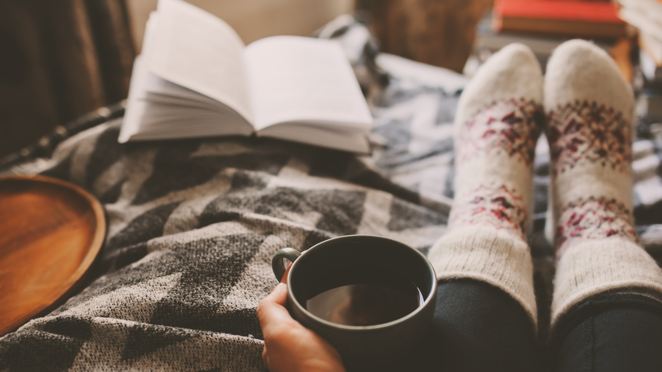 Person holding a cup of coffee with socks on and a book lying on a bed