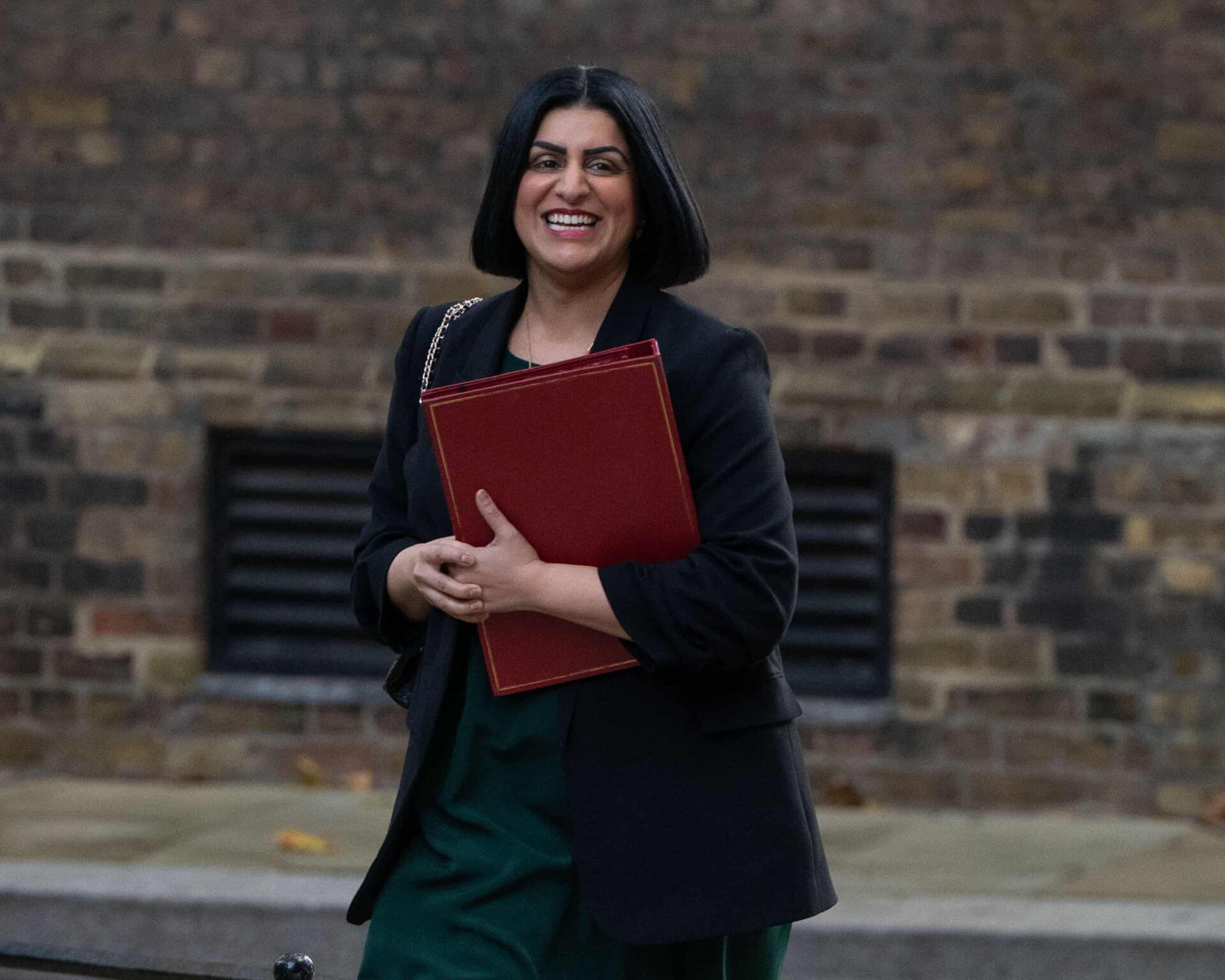 Shabana Mahmood, current Secretary of State for the UK Woman smiling in black coat holding a maroon file in her hands