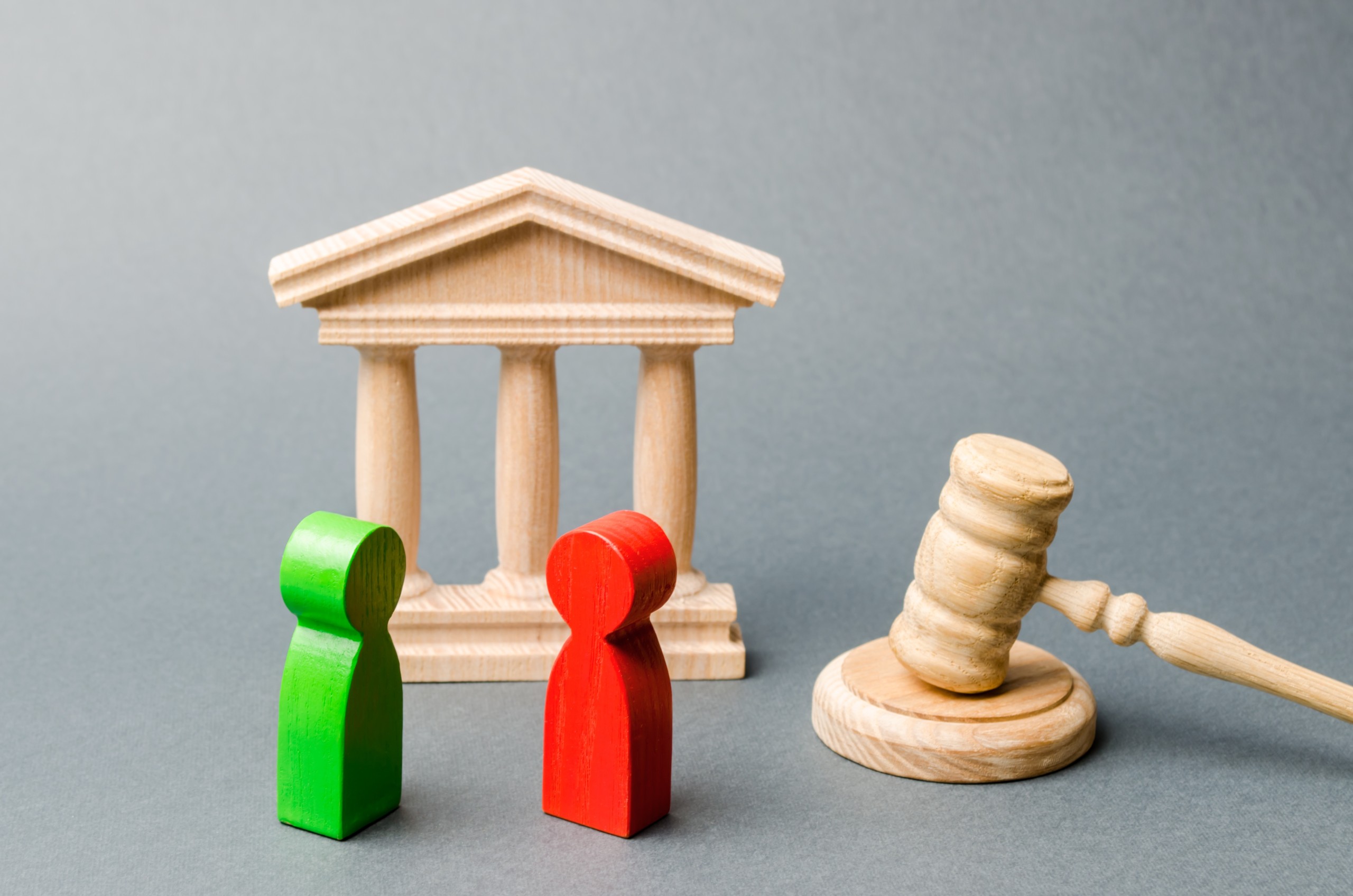 Two wooden figurines — one green, one red — standing before a miniature wooden courthouse, with a judge's gavel nearby, symbolizing a legal dispute.