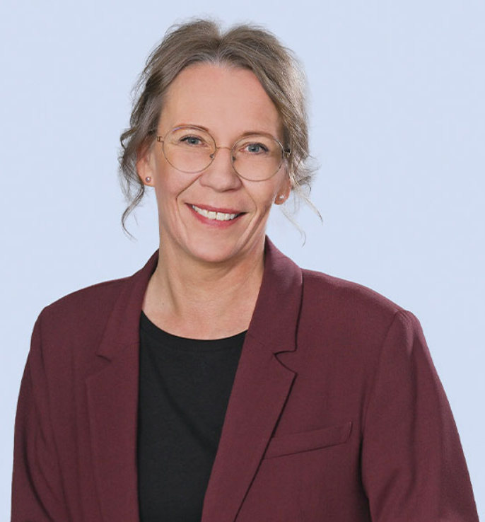 Headshot of Gitte Bjerremand Andersen, smiling, wearing round glasses and a dark red blazer against a light blue background.