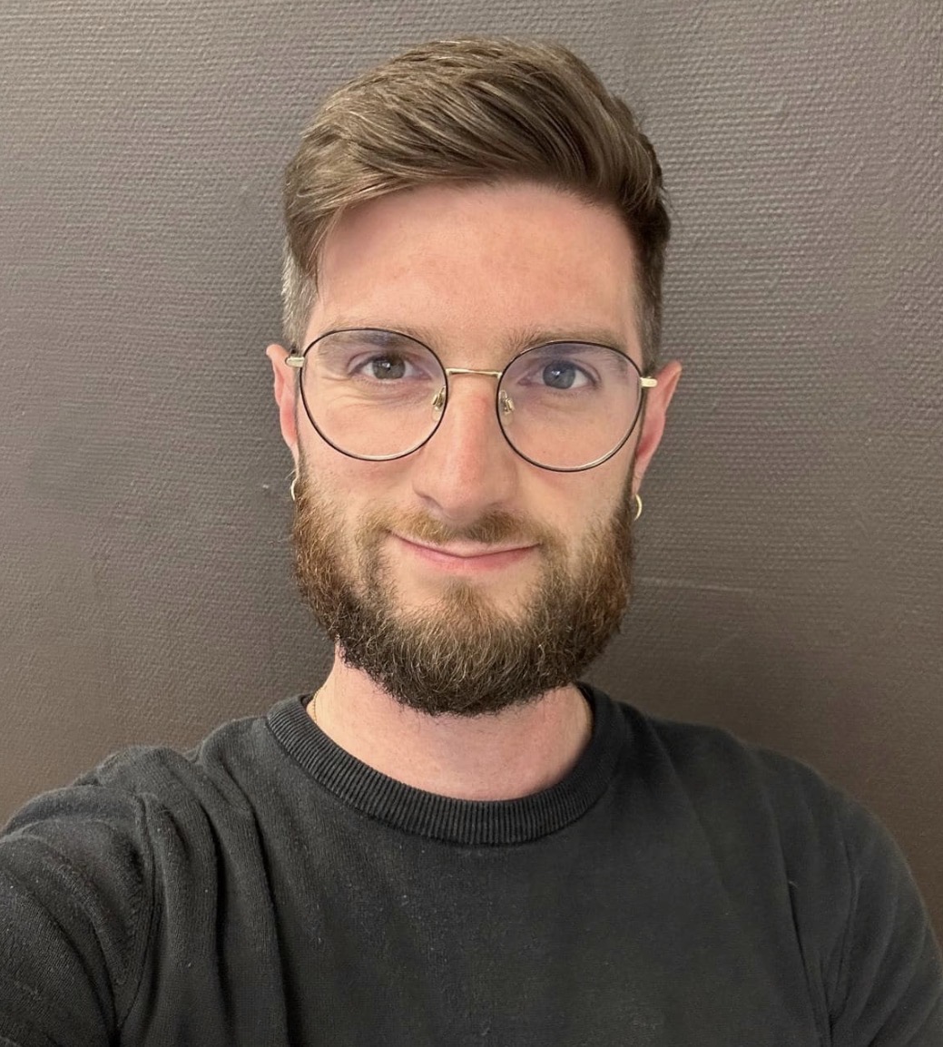 Headshot of Simone Giuseppe Uggeri, a bearded man with round glasses and a black sweater, against a grey textured background.