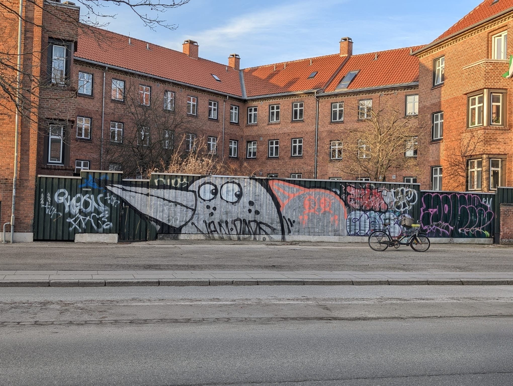 A construction hoarding covered in graffiti, including a large cartoon bird, in front of red-brick Copenhagen apartment buildings. A bicycle is parked alongside.