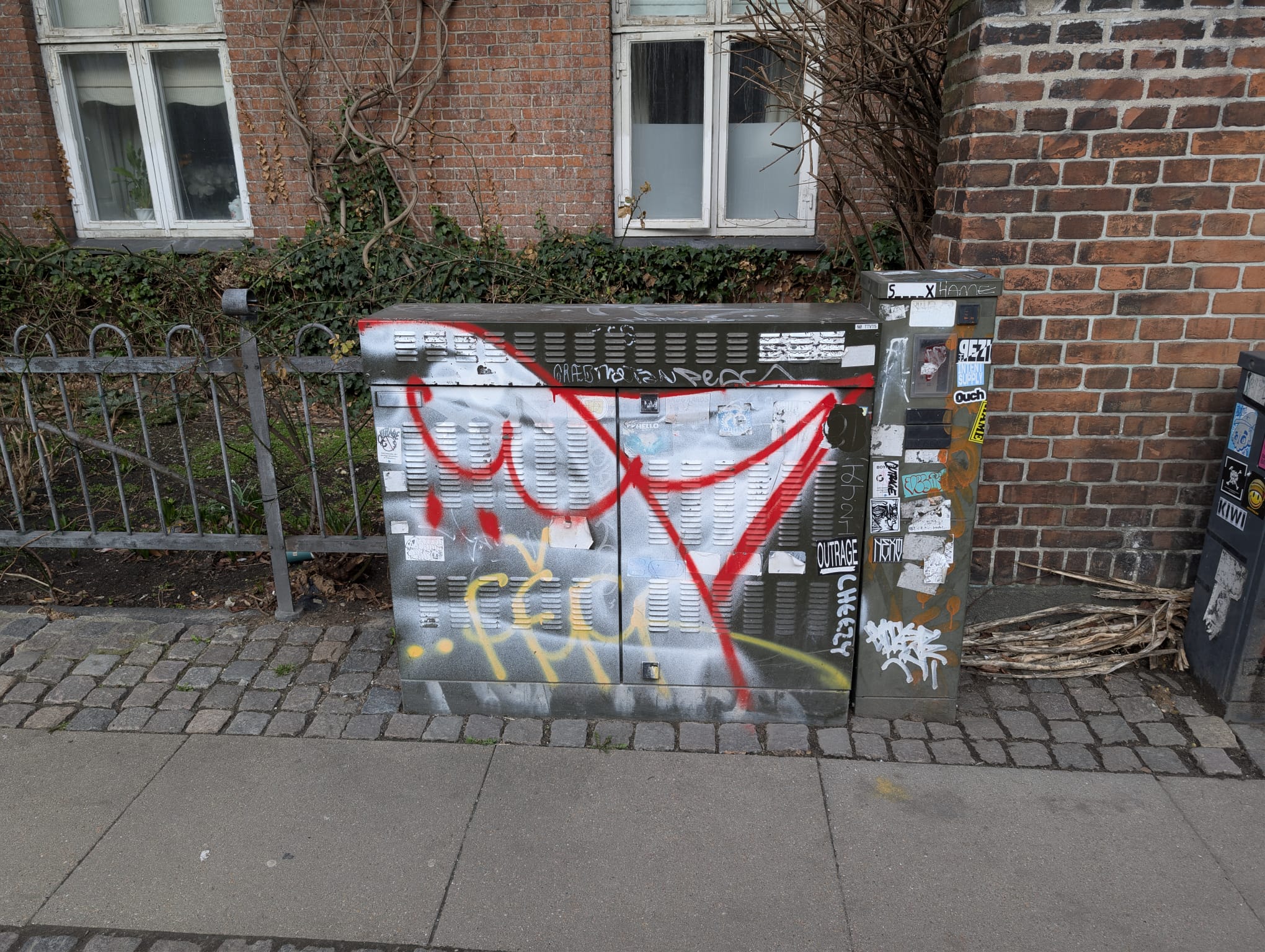 A street utility box covered in graffiti tags and stickers, including red spray-painted lines and yellow lettering, on a cobblestone pavement in front of a red-brick building in Copenhagen