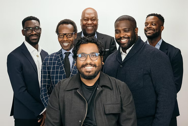 6 men in black suits on white background smiling at the camera