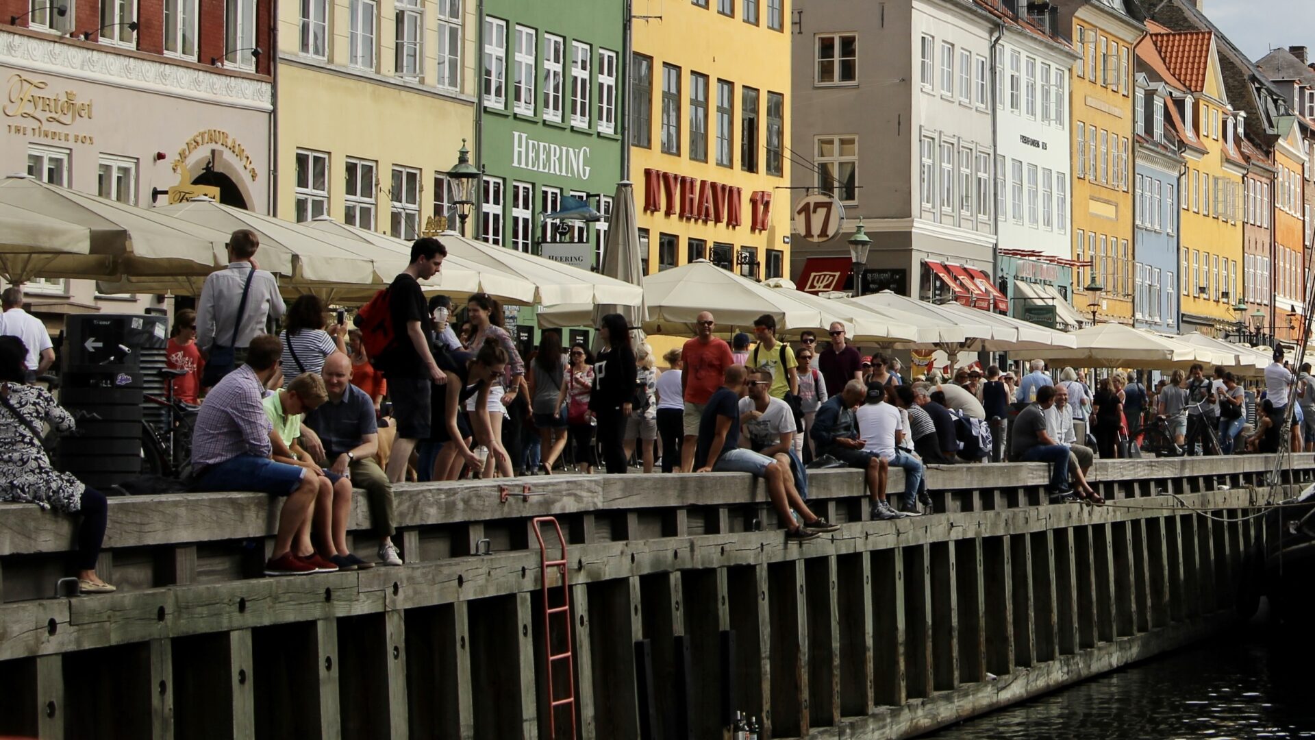 Crowded Nyhavn A dense crowd of tourists sitting and standing along the wooden quay at Nyhavn, with the canal's colourful 17th-century merchant houses and outdoor restaurant umbrellas stretching into the distance.