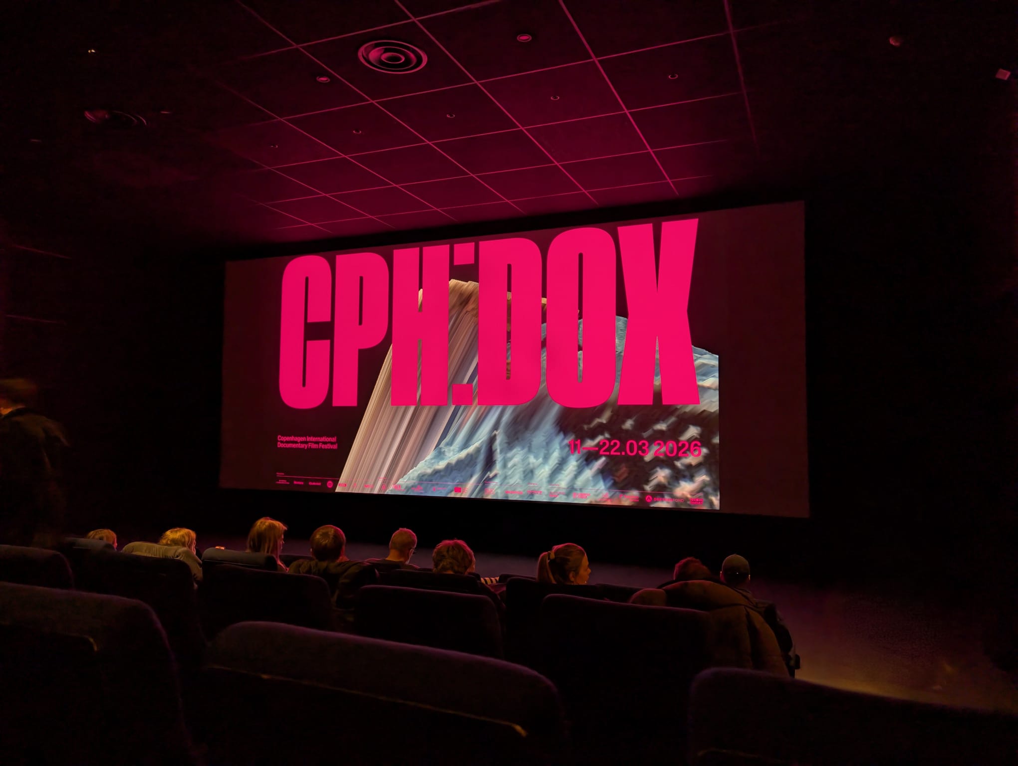 A darkened cinema audience facing a large screen displaying the bold pink CPH:DOX logo with the dates 11–22 March 2026.