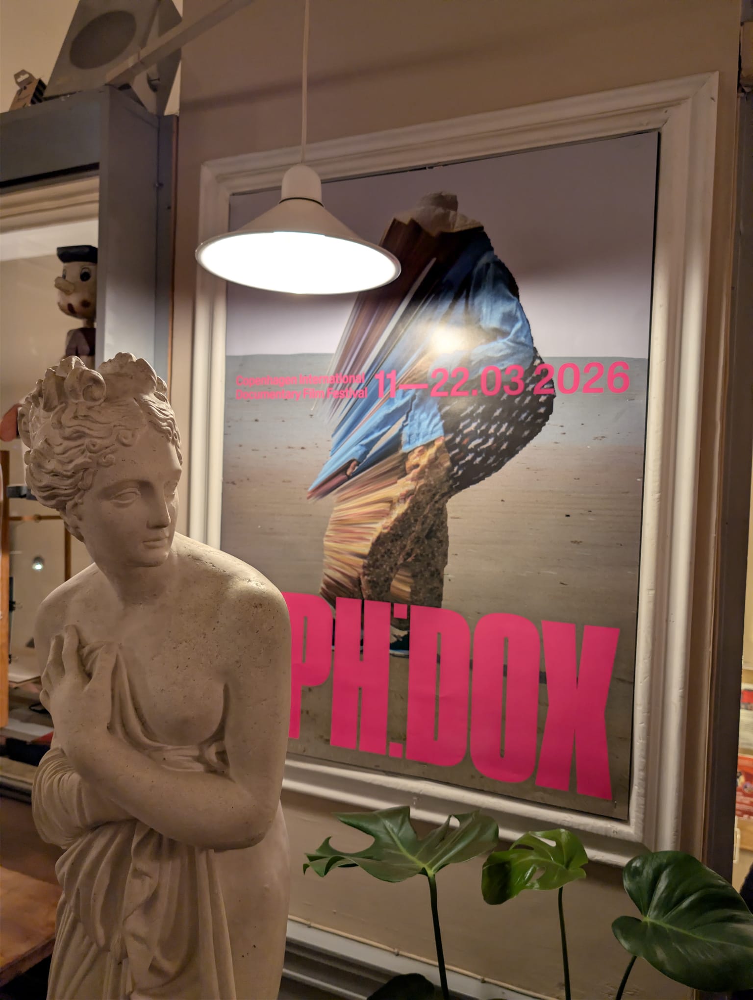 A classical white female bust sculpture in the foreground, with a framed CPH:DOX 2026 festival poster hanging on the wall behind it, lit by a pendant lamp.