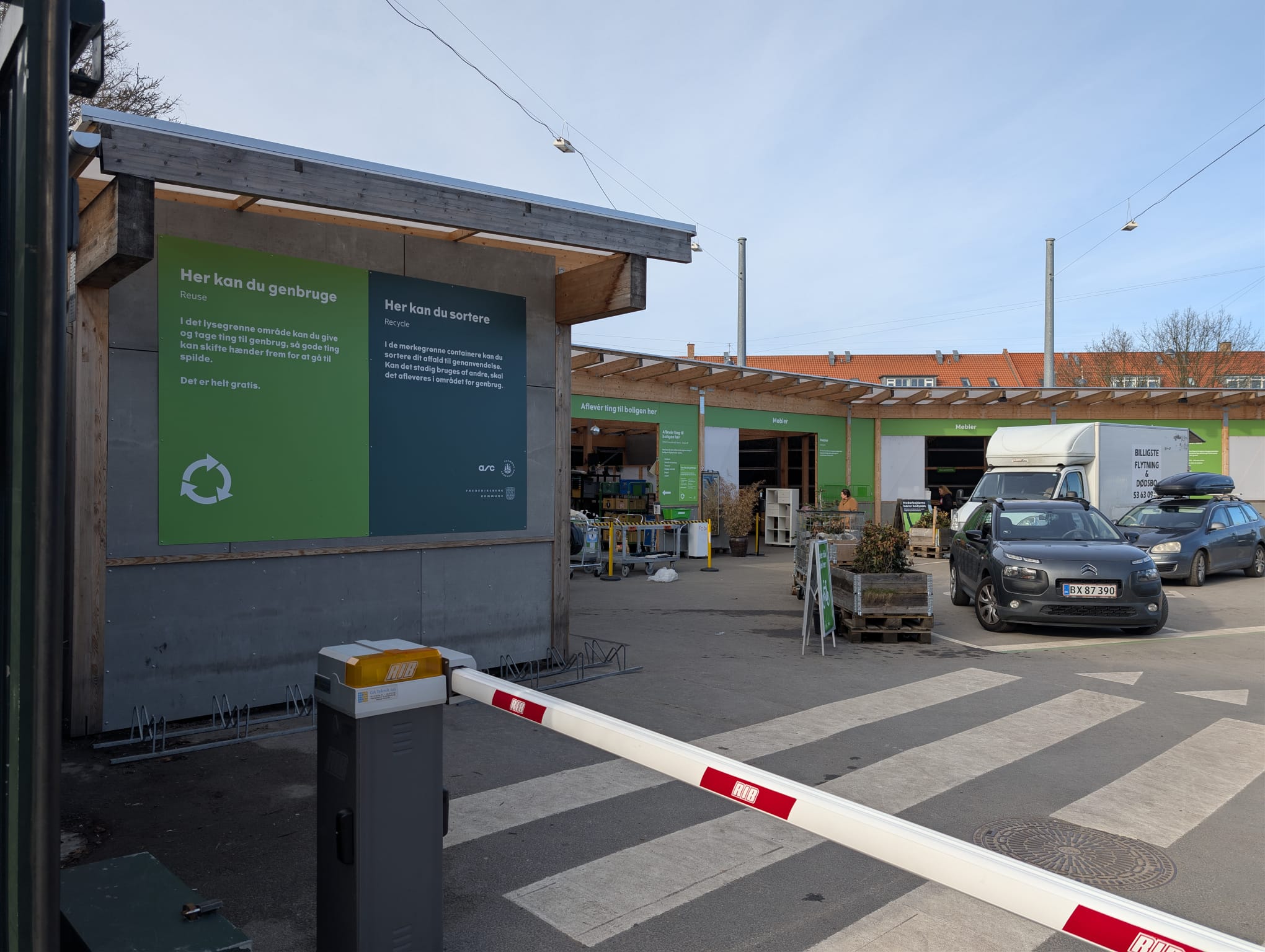 Entrance to a Danish recycling centre (genbrugsplads) with a barrier gate and green information signs explaining reuse and sorting options. Cars and a truck are visible in the background by the drop-off area.