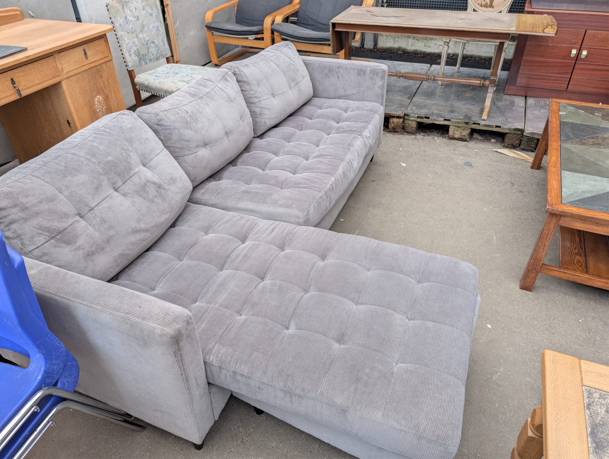 A large grey tufted corner sofa displayed outdoors among other second-hand furniture at a reuse centre, surrounded by chairs, tables, and wooden cabinets.