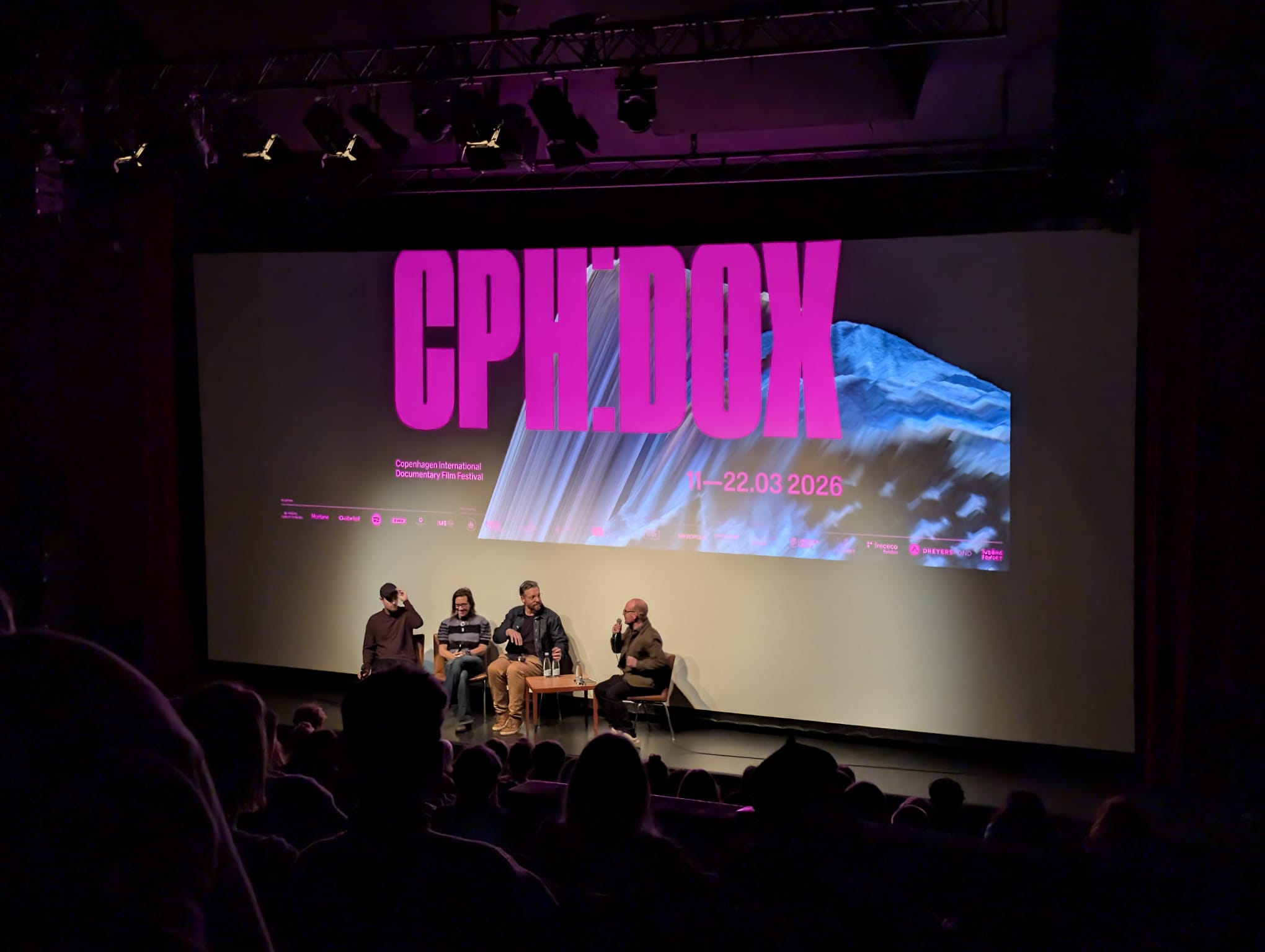 Three panelists seated on stage in front of a large CPH:DOX screen, speaking to a full cinema audience in a darkened theatre