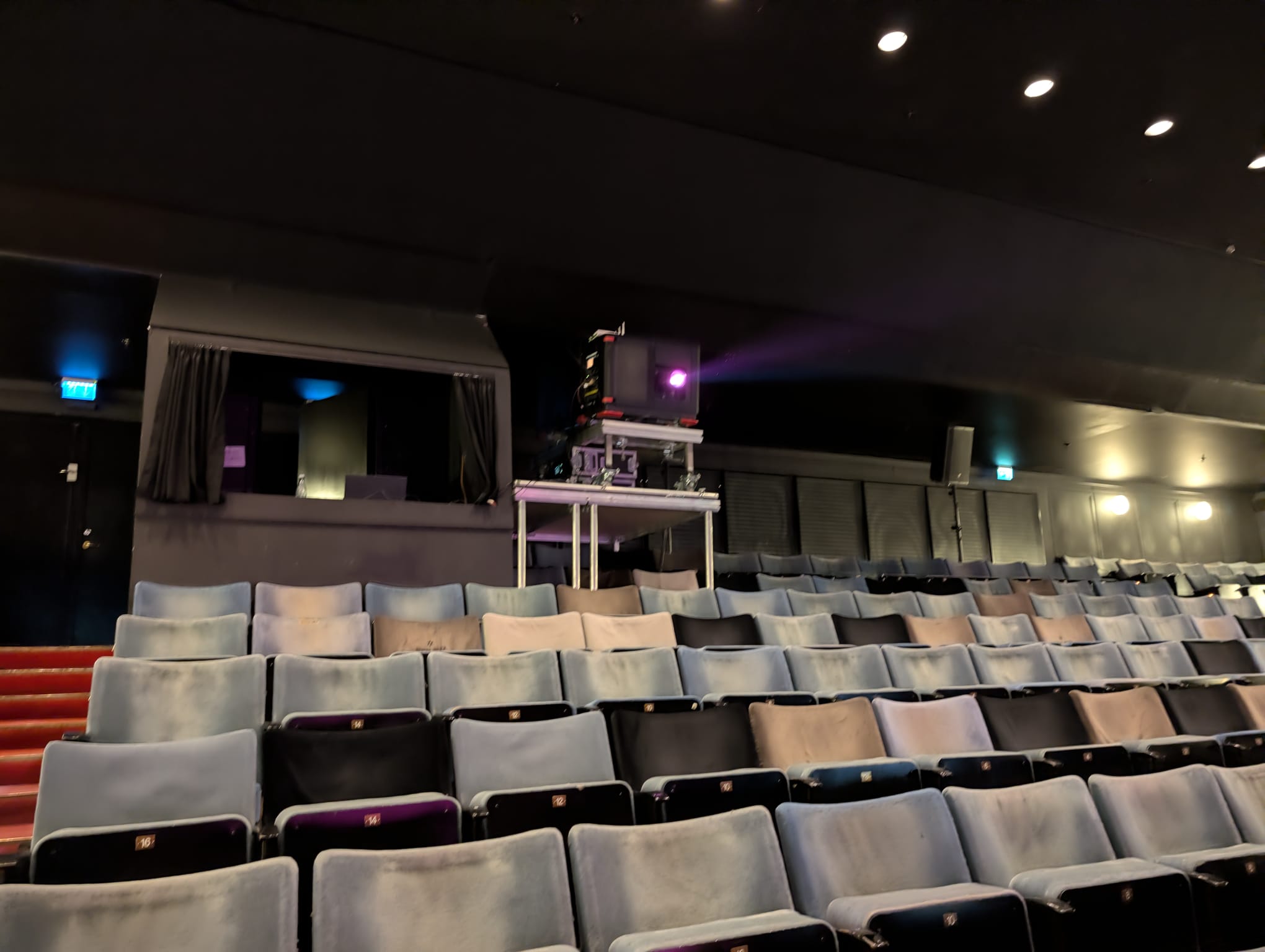 Rows of empty grey velvet cinema seats viewed from a mid-row angle, with a film projector glowing purple in the background