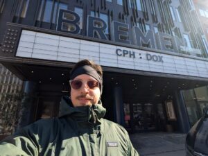 Selfie outside Bremen Cinema A person in sunglasses and a green jacket takes a selfie outside the Bremen cinema, whose marquee reads 'CPH:DOX'.