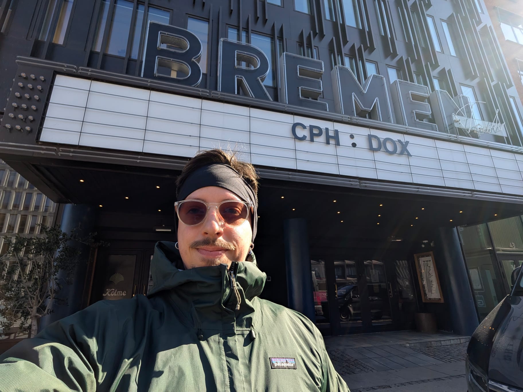 A person in sunglasses and a green jacket takes a selfie outside the Bremen cinema, whose marquee reads 'CPH:DOX'.