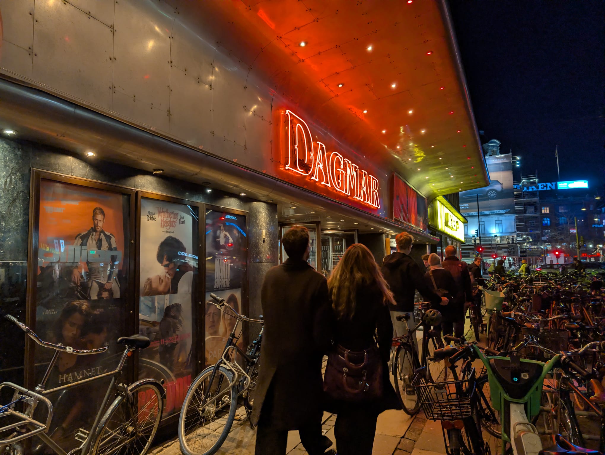The Dagmar cinema at night in Copenhagen, with its glowing red neon sign, film posters in the windows, and a crowd of people and parked bicycles on the pavement outside.