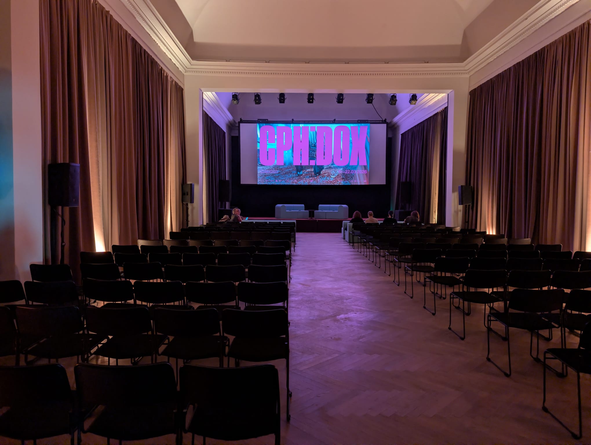 A large, elegantly draped event hall with rows of black chairs facing a stage where a few people are seated, with the CPH:DOX logo displayed on a wide screen in warm pink-purple lighting.