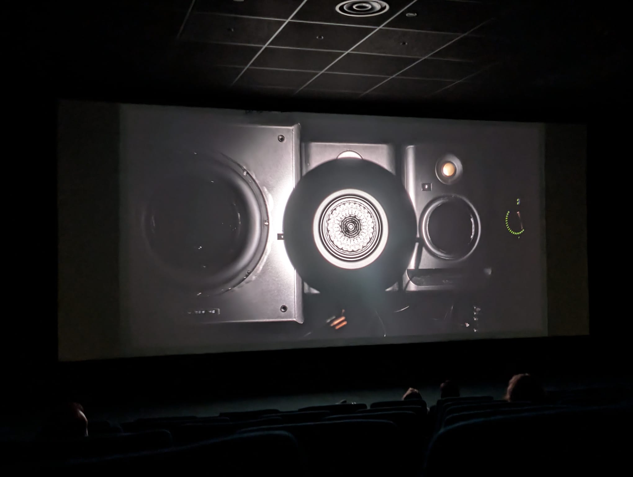 A cinema screen showing a close-up shot of a professional studio monitor speaker with a glowing tweeter, viewed from the darkened audience.