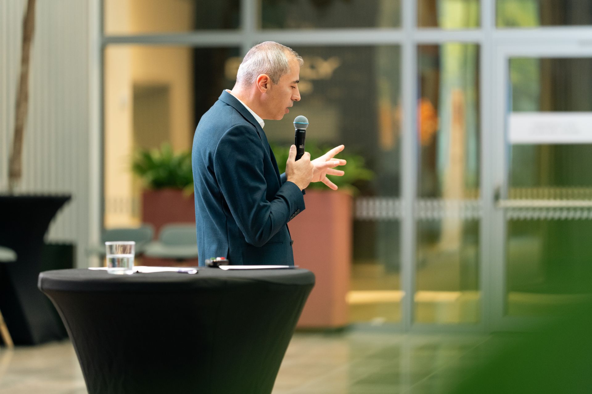 Florin Lungu stands at a high cocktail table holding a microphone and gesturing with one hand, speaking at an indoor event with large glass windows and plants in the background.