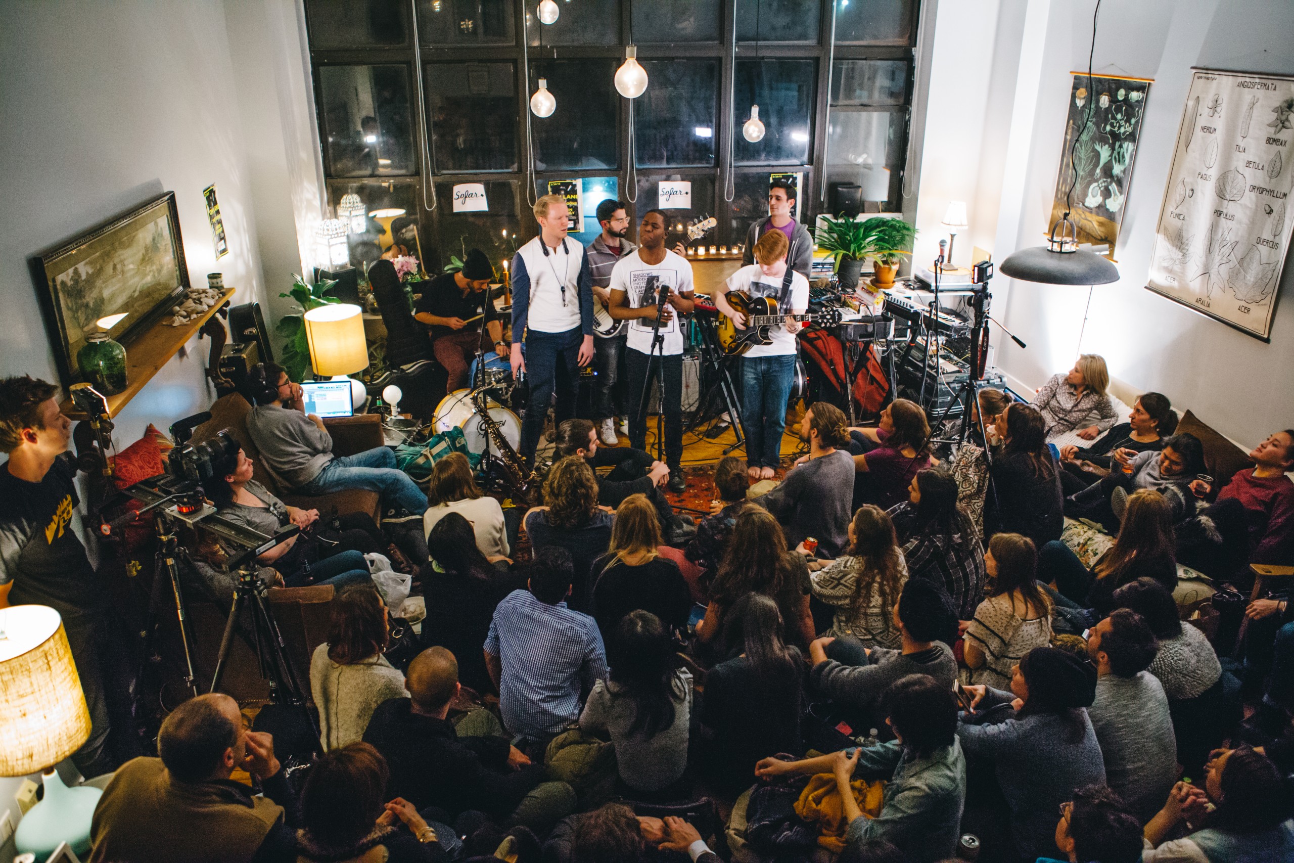 A band performs at an intimate Sofar Sounds concert in a warmly lit apartment, surrounded by a large crowd sitting on the floor and every available surface.