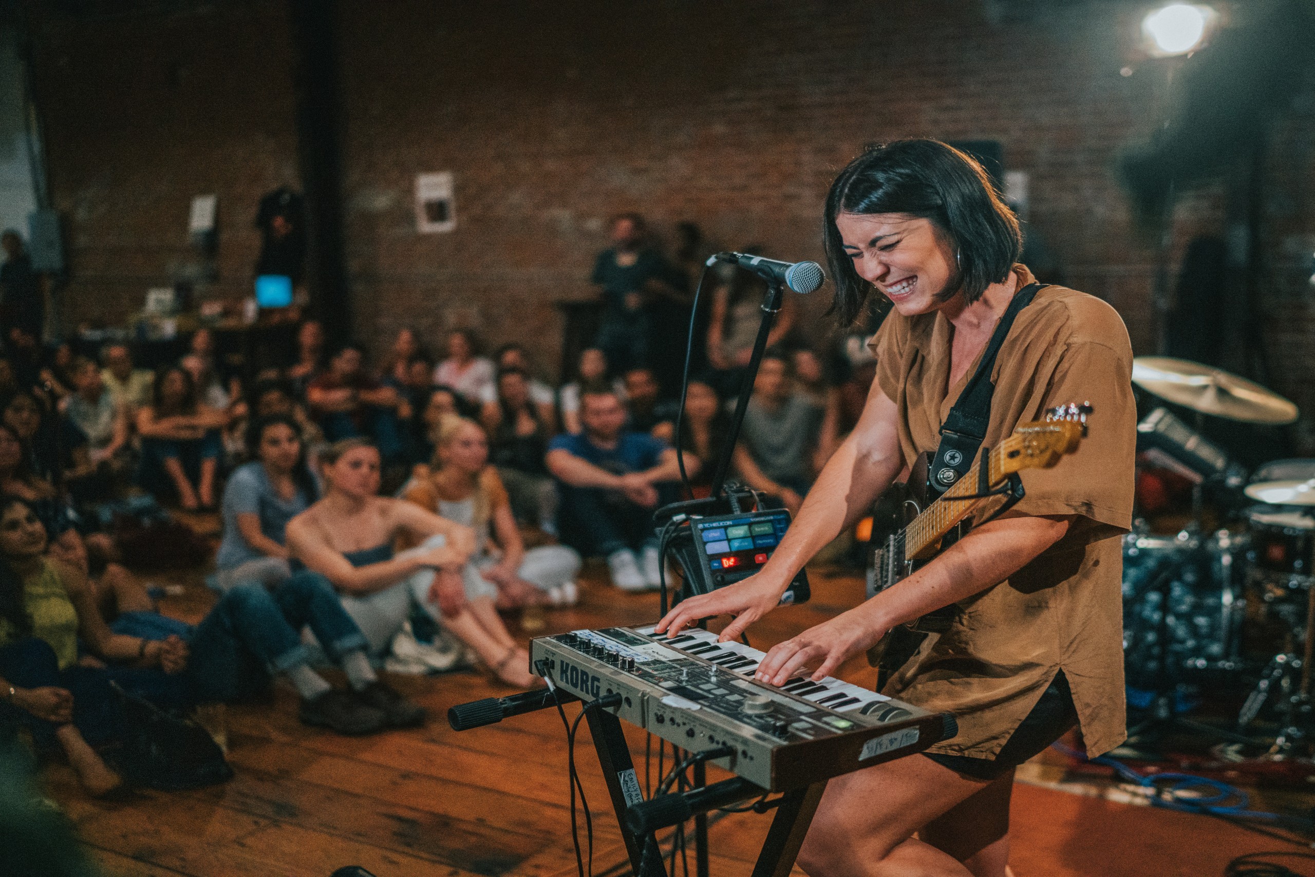 A smiling woman with a guitar slung over her shoulder plays a Korg keyboard at a Sofar Sounds show, with an engaged seated audience filling a brick-walled room behind her.