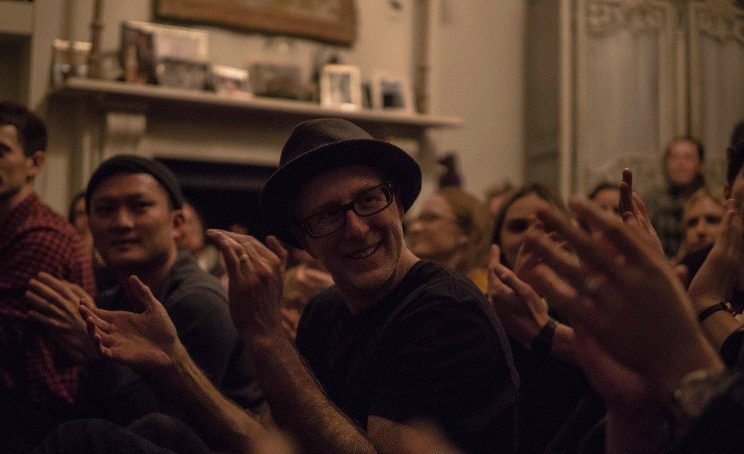 A crowd of audience members clapping in a warmly lit living room, with a smiling man in a black fedora and glasses at the centre.