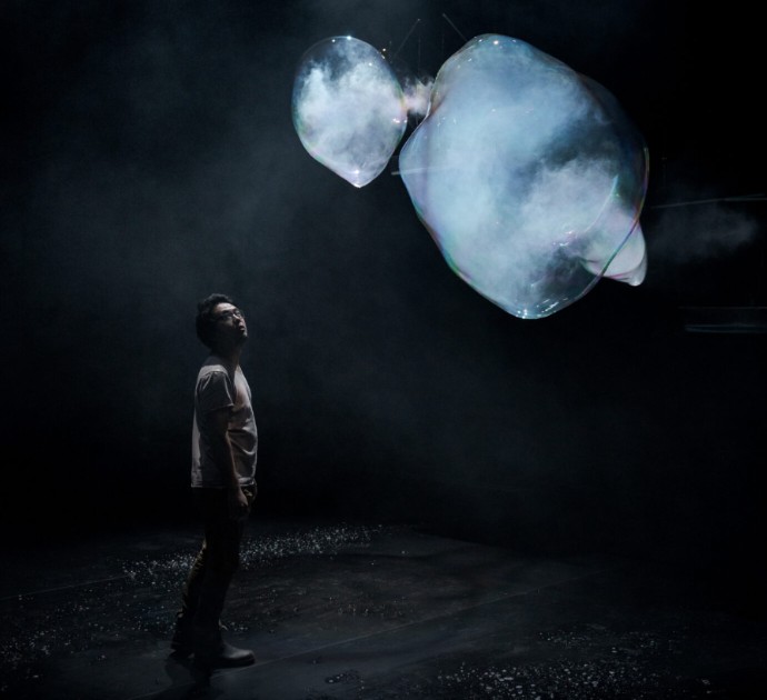 A person gazes up at large iridescent soap bubbles filled with mist floating in a dark installation space