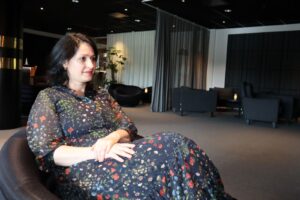 Florina Lungu A woman in a dark floral dress sits in an armchair with hands clasped, looking thoughtfully off to the side, in a softly lit lounge area with grey curtains and dark furniture