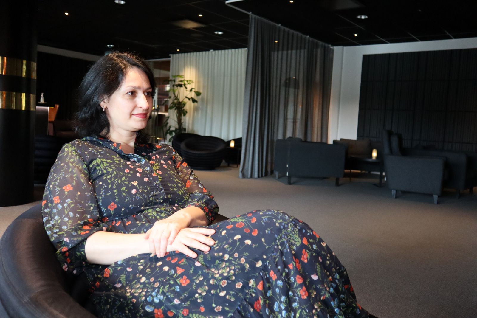 Florina Lungu A woman in a dark floral dress sits in an armchair with hands clasped, looking thoughtfully off to the side, in a softly lit lounge area with grey curtains and dark furniture