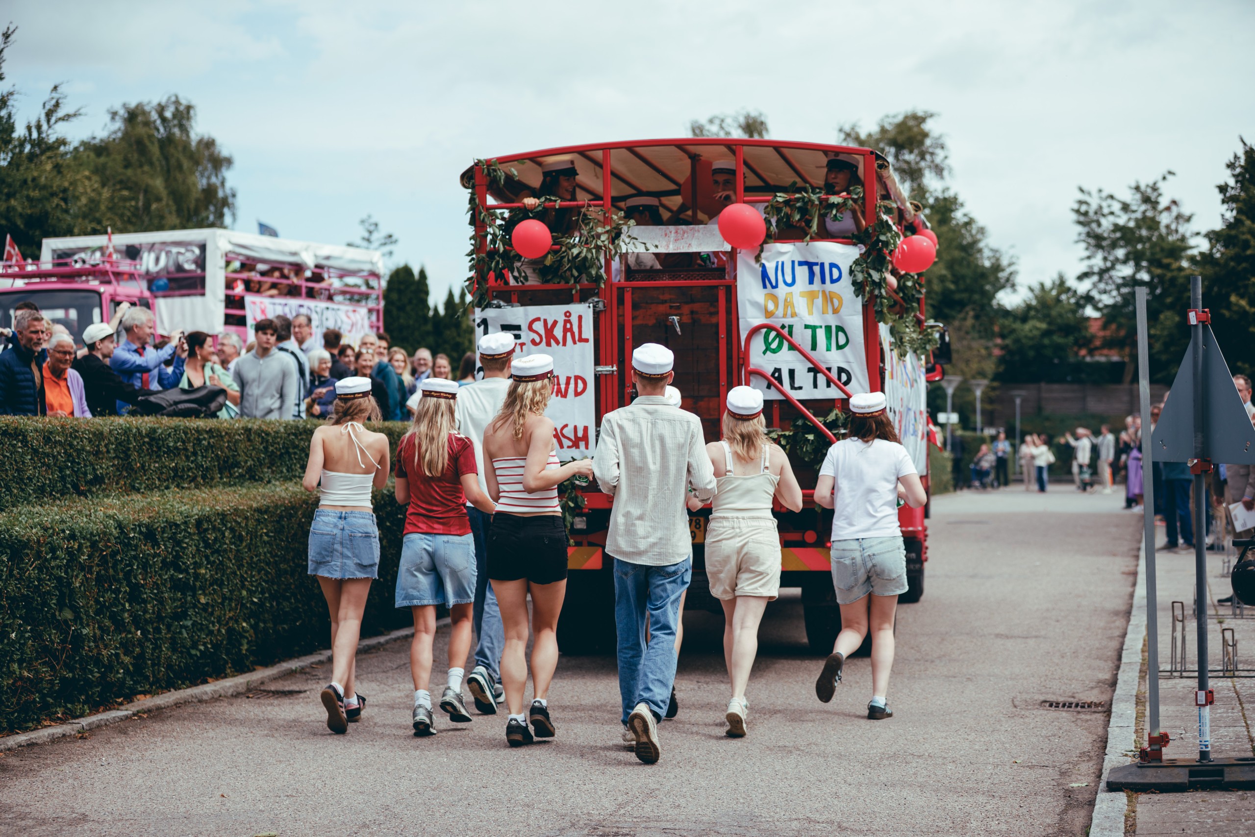 A group of Danish graduates in white student caps walk behind a red decorated graduation truck (studenterkørsel) covered in hand-painted banners, balloons, and greenery, as a crowd watches from behind a hedge