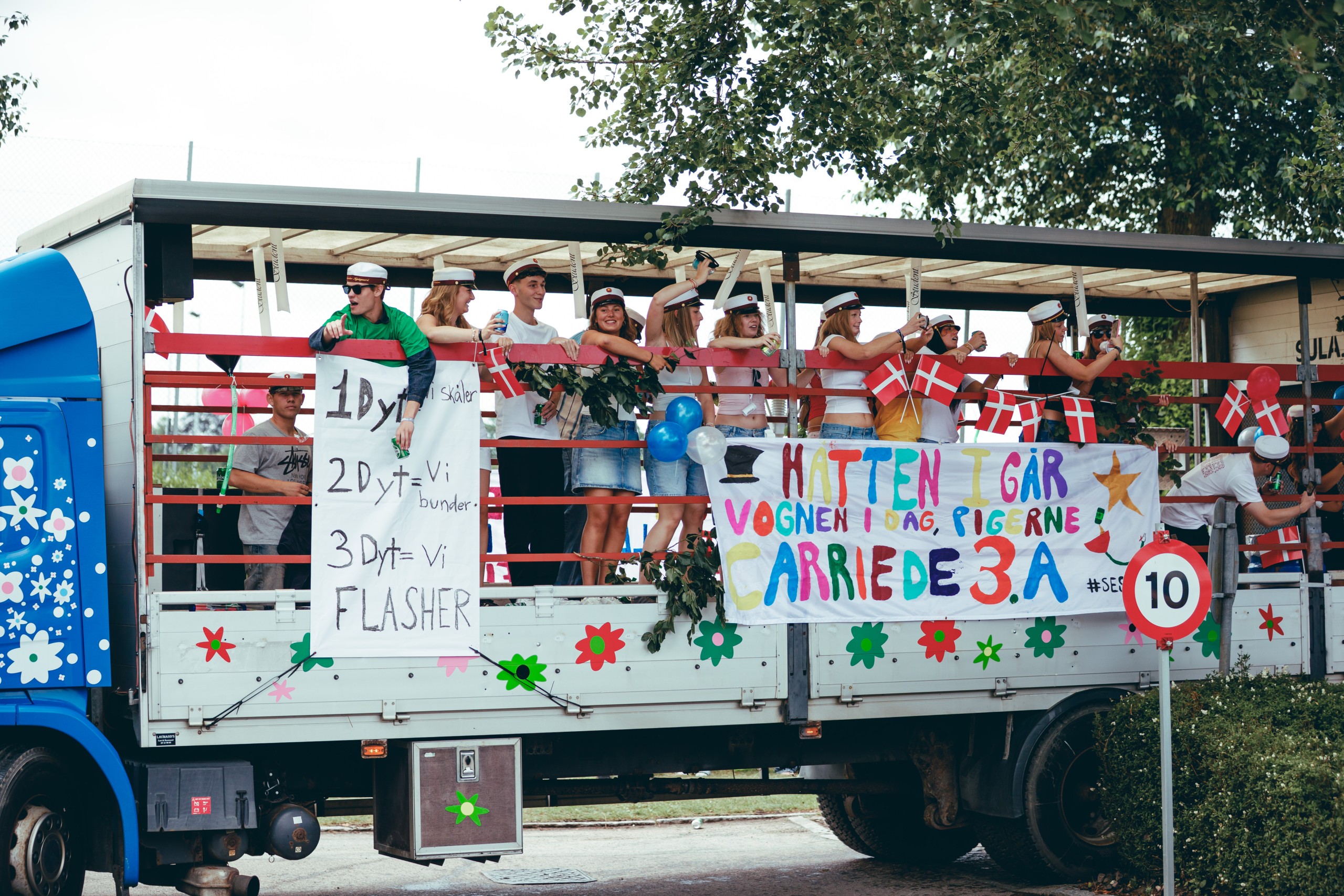 A group of Danish students in caps and sashes stand on a flower-decorated flatbed truck, holding Danish flags and colourful hand-painted banners with jokes, as part of a graduation parade (studenterkørsel).