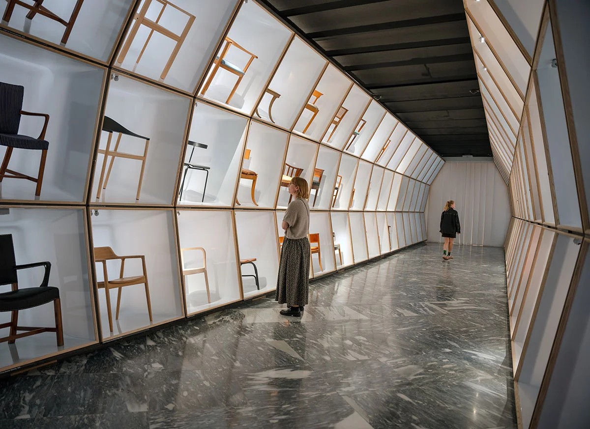 Two visitors walk a long corridor lined with individually lit display cases showing Danish design chairs at Designmuseum Danmark