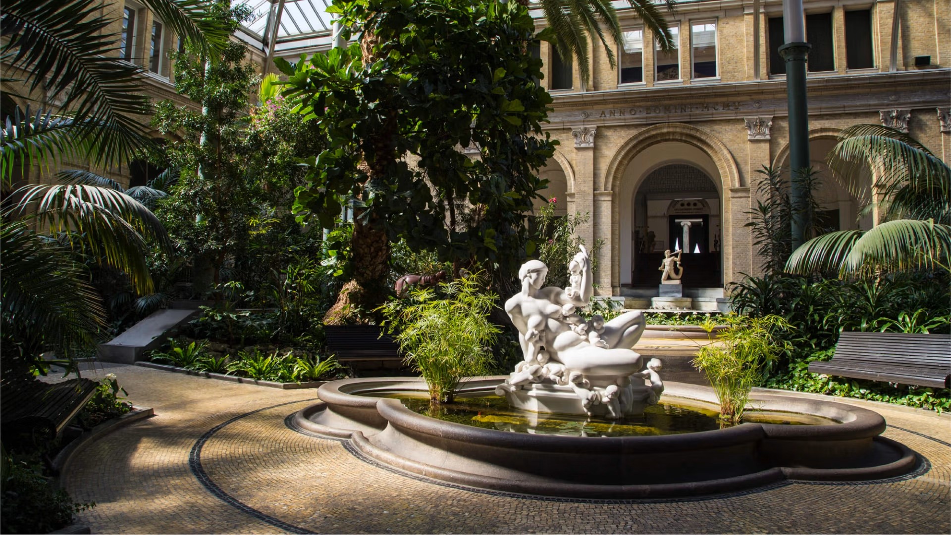 A sunlit indoor garden with tropical plants, a marble fountain sculpture, and a neoclassical arcade at the Ny Carlsberg Glyptotek.