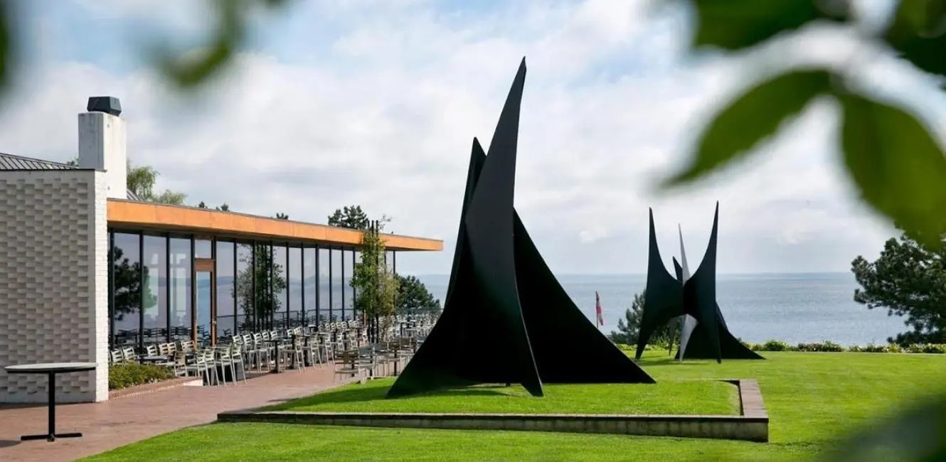 Two large black steel Calder stabile sculptures on the Louisiana Museum lawn overlooking the sea, with the museum café terrace behind.