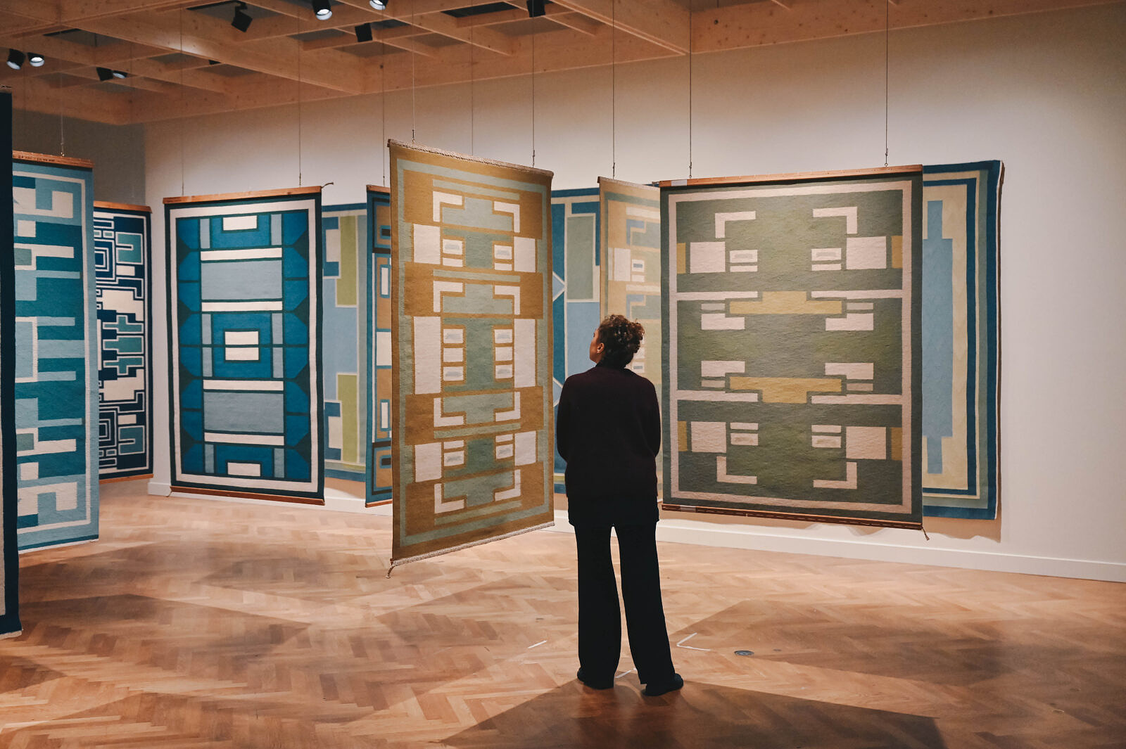 A visitor stands before a row of large geometric woven tapestries in blue, gold, and green tones suspended from the ceiling at SMK.