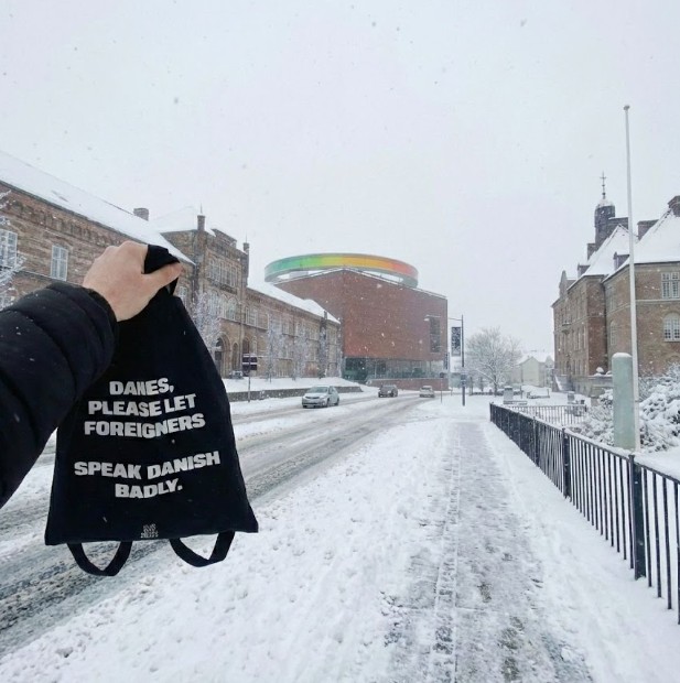 A hand holds up a black tote bag reading 'Danes, please let foreigners speak Danish badly' against a snowy street scene, with the colourful circular ARoS museum rooftop visible in the background.