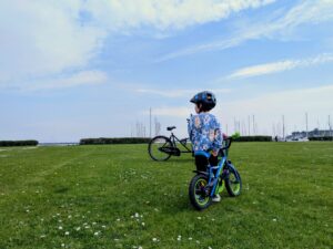 Taking a break by the marina on a family bike ride Child in a helmet standing with a small blue bike on a grassy lawn beside a marina full of sailboats