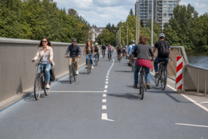 Cyclists crossing a dedicated bike bridge A busy two-lane cycle bridge with cyclists riding in both directions, flanked by trees and a river, with urban buildings visible in the background.