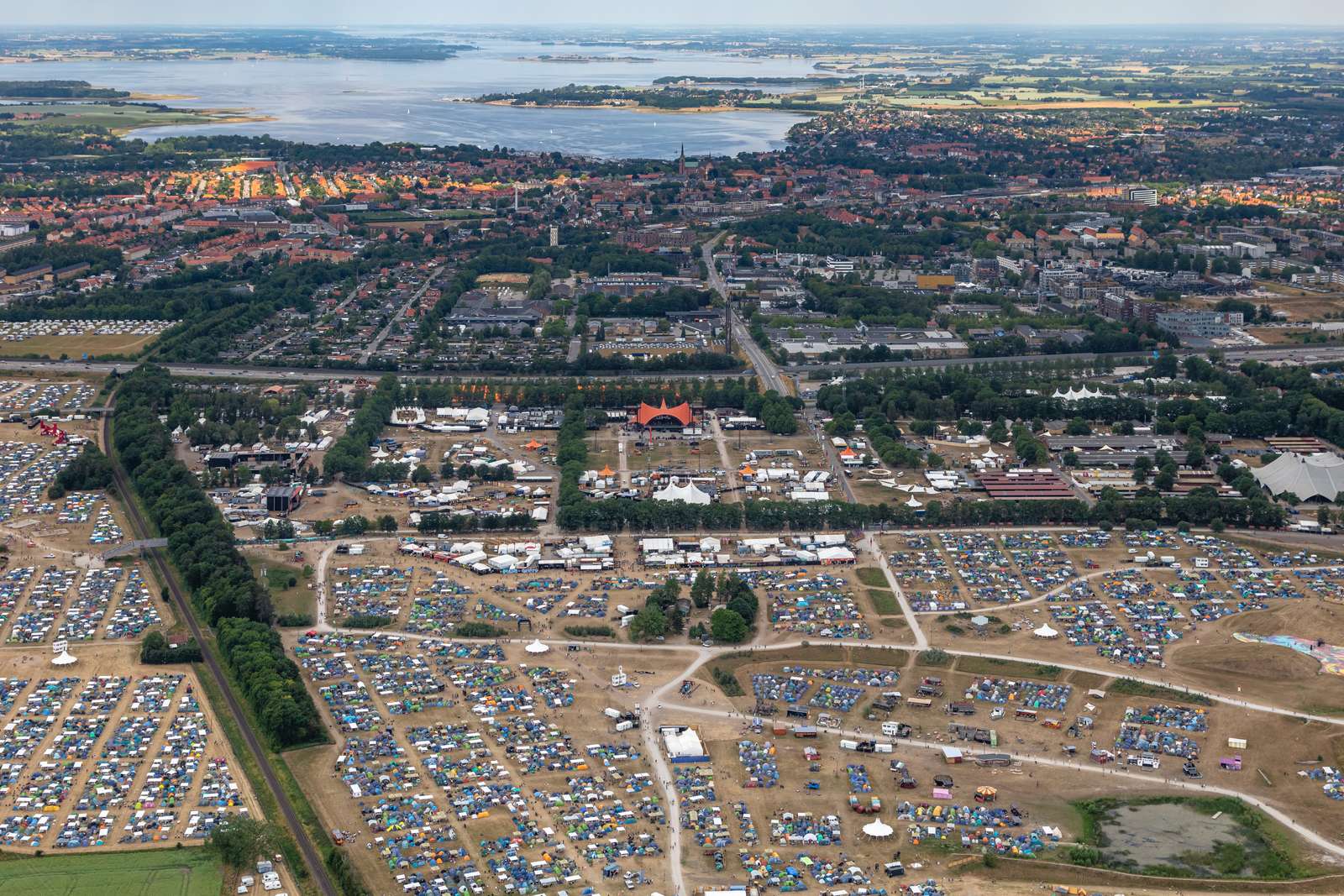 Aerial shot of Roskilde Festival with thousands of colourful tents spreading across the foreground, the iconic orange stage visible in the middle, and the town of Roskilde and its fjord stretching into the distance