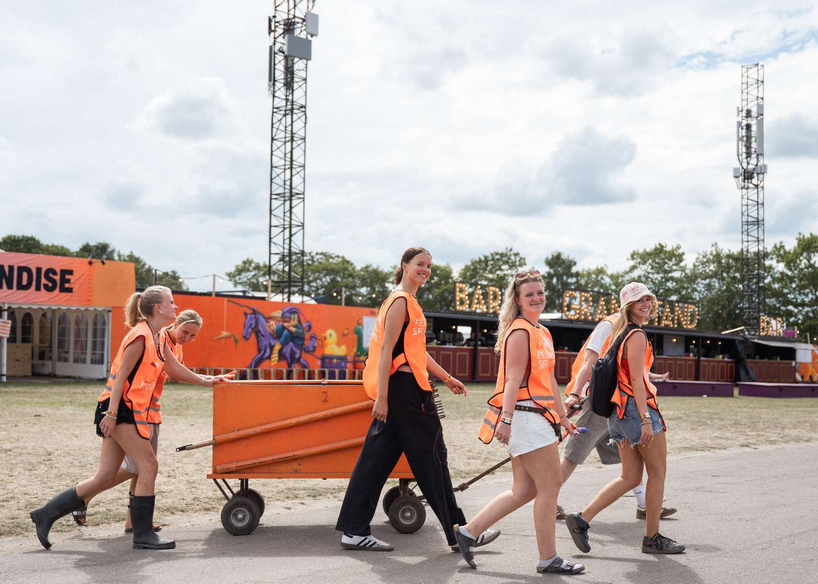 Four smiling Roskilde Festival volunteers in orange high-visibility vests push a large orange equipment cart across the open festival grounds, with the Bar Grand Stand visible behind them.