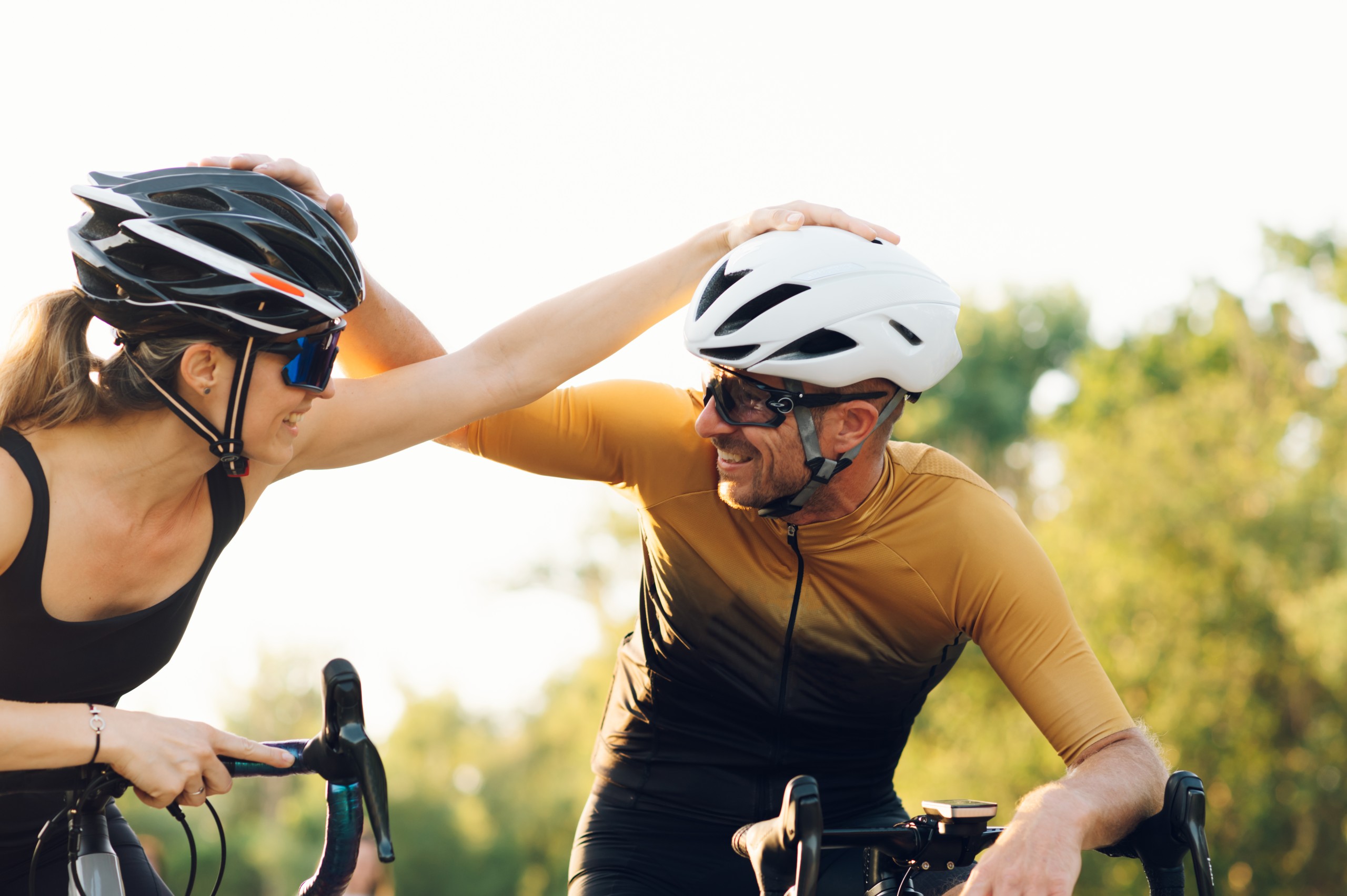 Two road cyclists in helmets and sunglasses playfully tap each other's helmets, smiling, against a warm backlit outdoor background