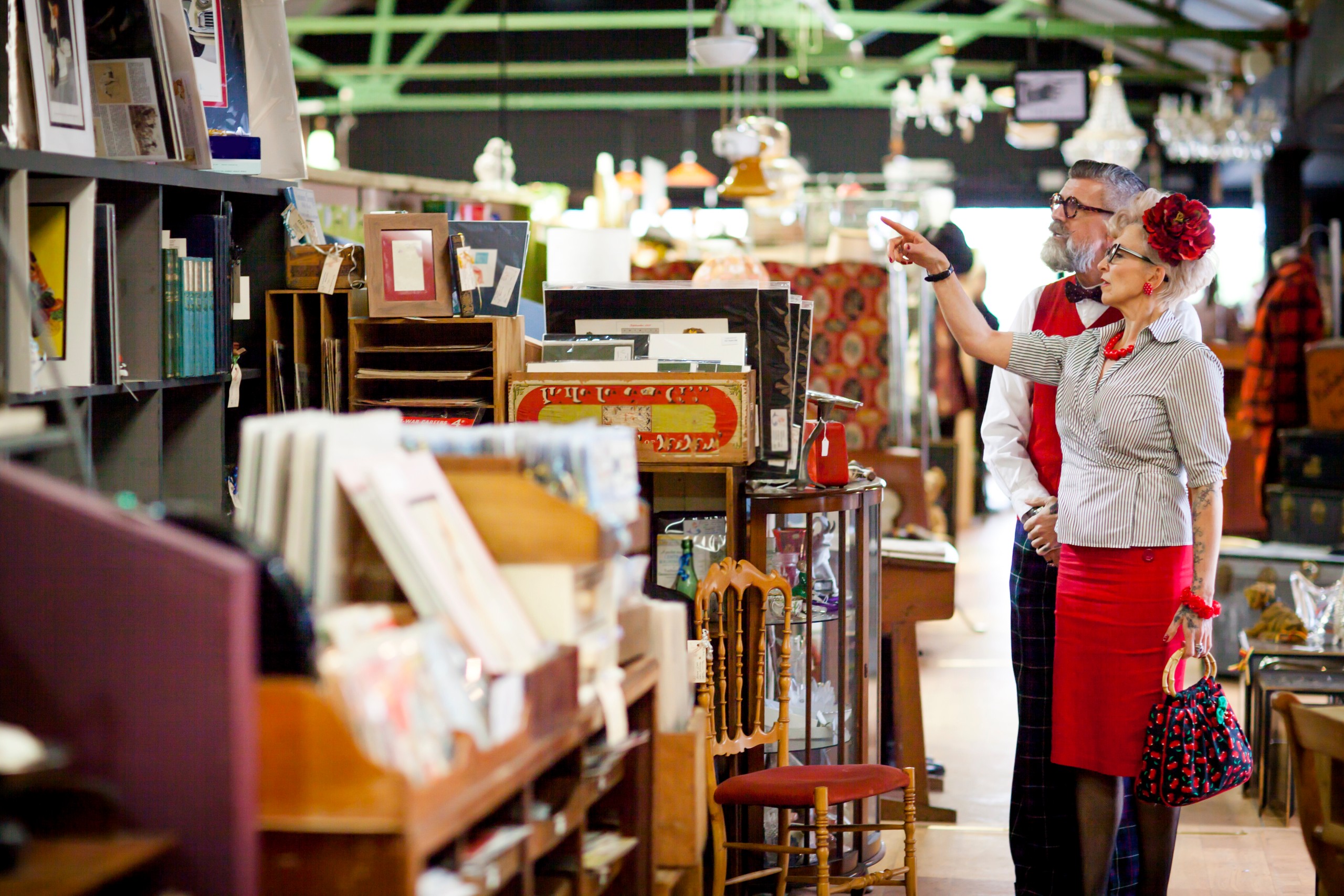 Vintage-styled couple browsing framed prints and antique furniture at an indoor antiques market