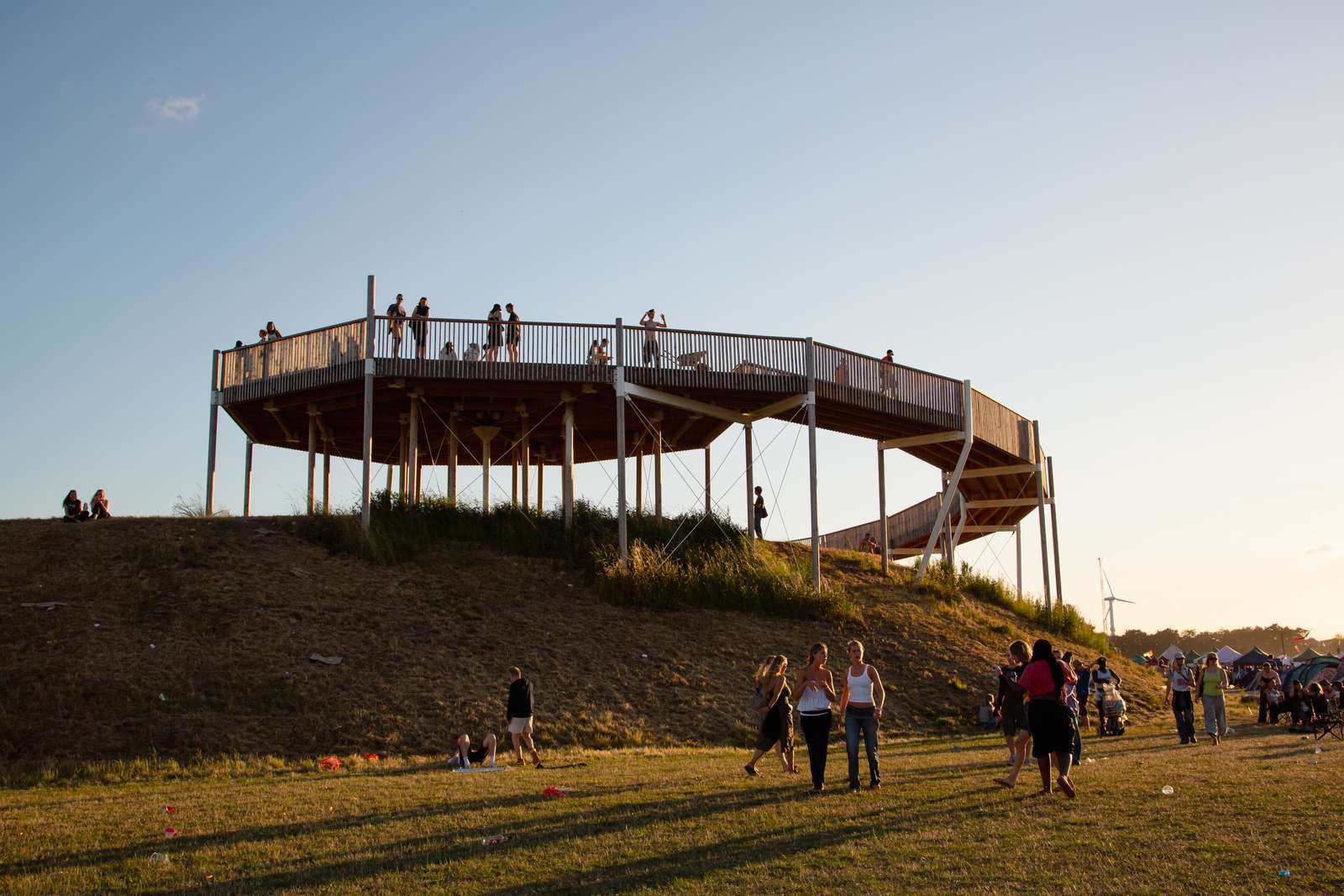 Festival-goers stand on a raised circular wooden viewing platform on a hilltop at golden hour, with others milling below and a wind turbine visible on the horizon