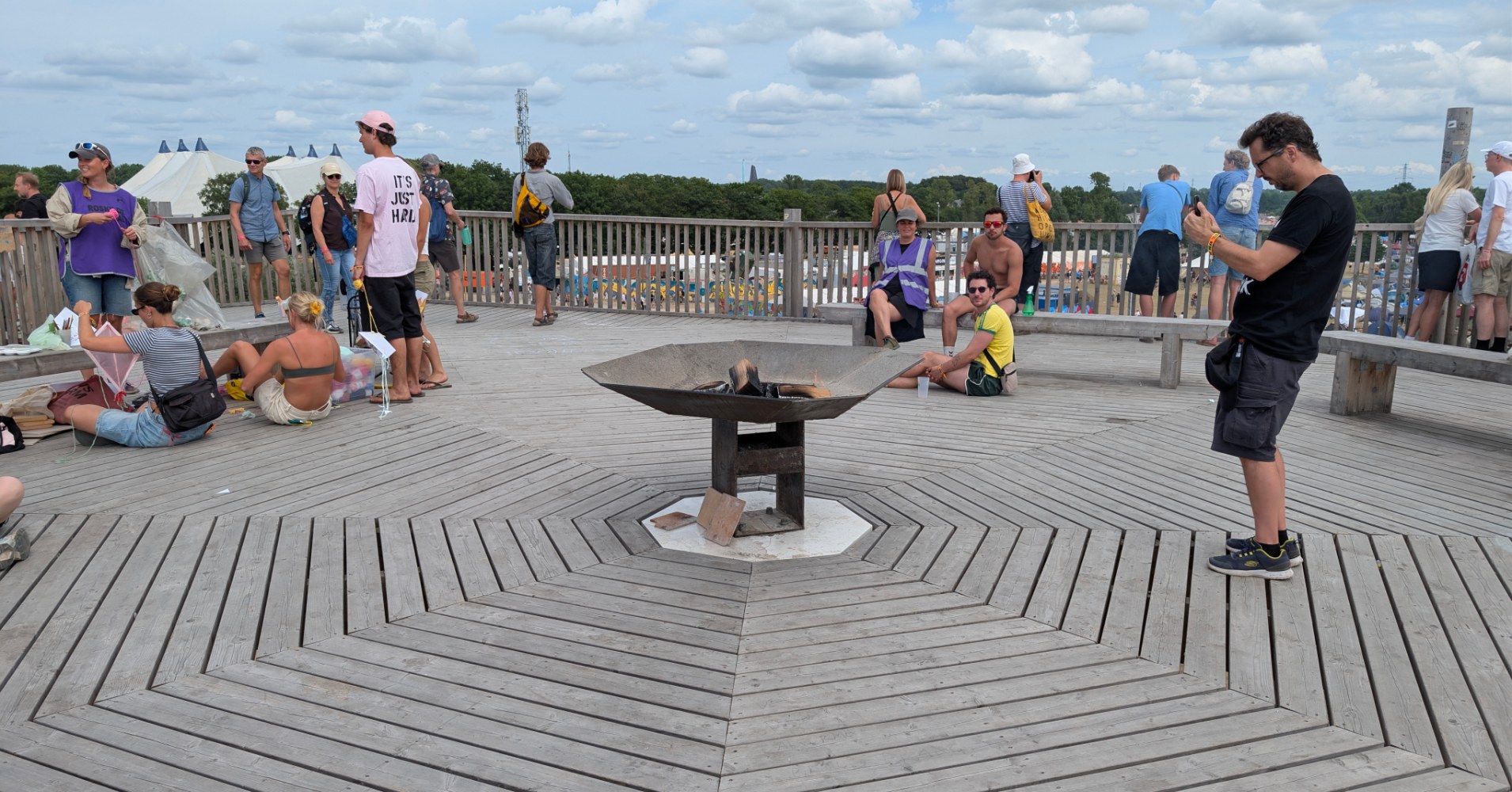 Festival visitors relax around a central fire bowl on the wooden hilltop platform, with the campsite and festival grounds visible beyond the railings.