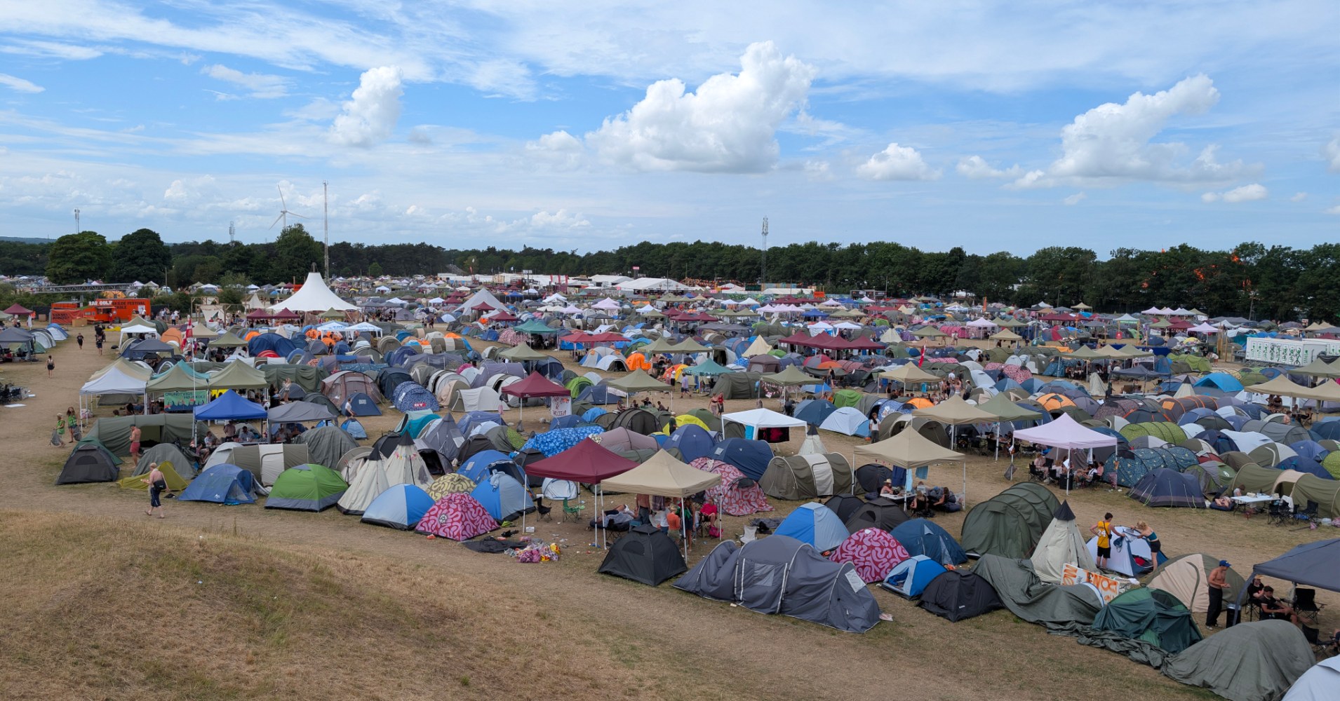 Hundreds of colourful tents and gazebos packed tightly across a dry festival campsite field under a partly cloudy summer sky.