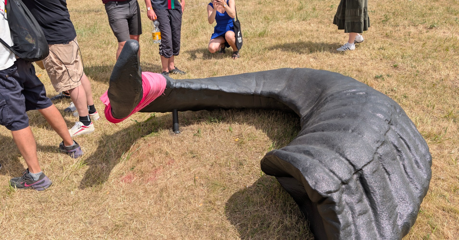Festival visitors interact with a large curved black sculptural form lying on the dry grass, with one person climbing inside it while others photograph.