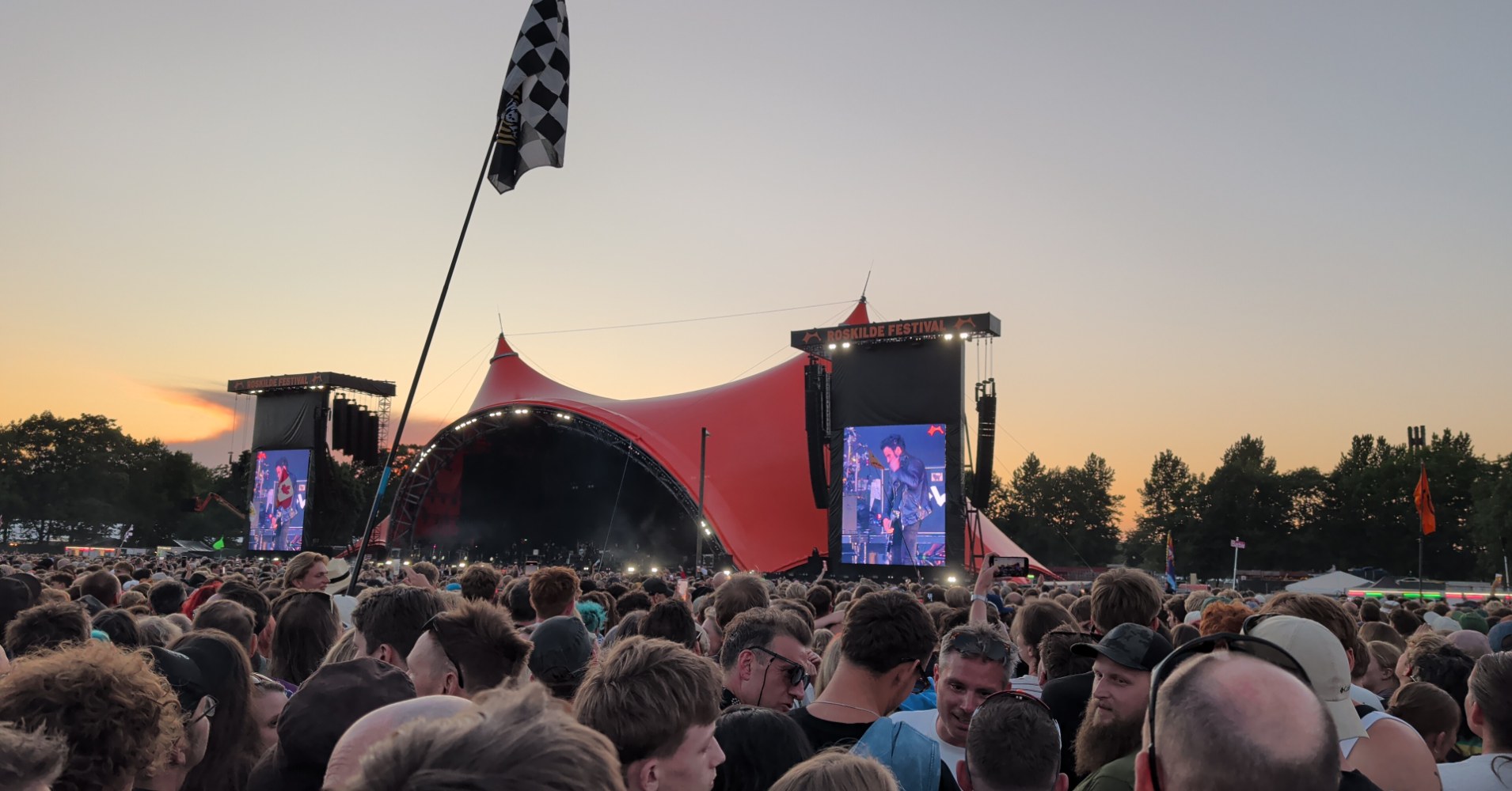 A dense crowd faces the iconic red-roofed Orange Stage at Roskilde Festival during a sunset performance, with large video screens showing the artist and a chequered flag waving above the crowd.