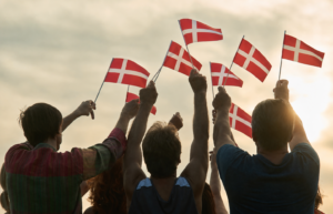 Group waving Danish flags at sunset Silhouettes of a group of people seen from behind, raising their arms and waving small Danish flags against a warm, cloudy sunset sky