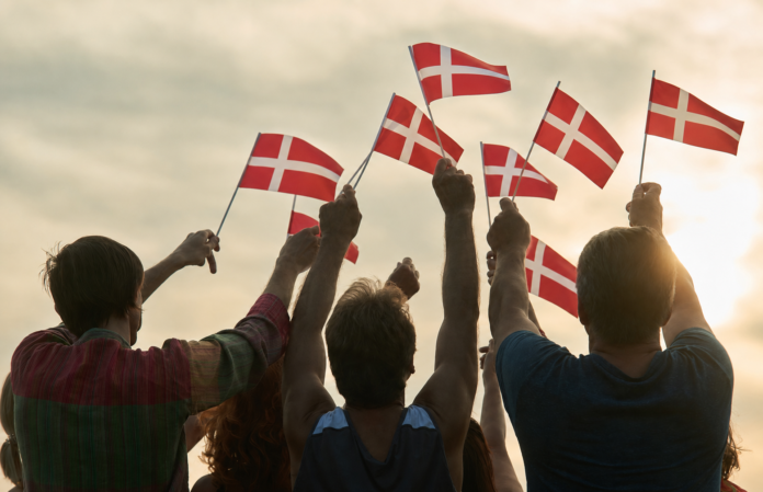 Group waving Danish flags at sunset Silhouettes of a group of people seen from behind, raising their arms and waving small Danish flags against a warm, cloudy sunset sky