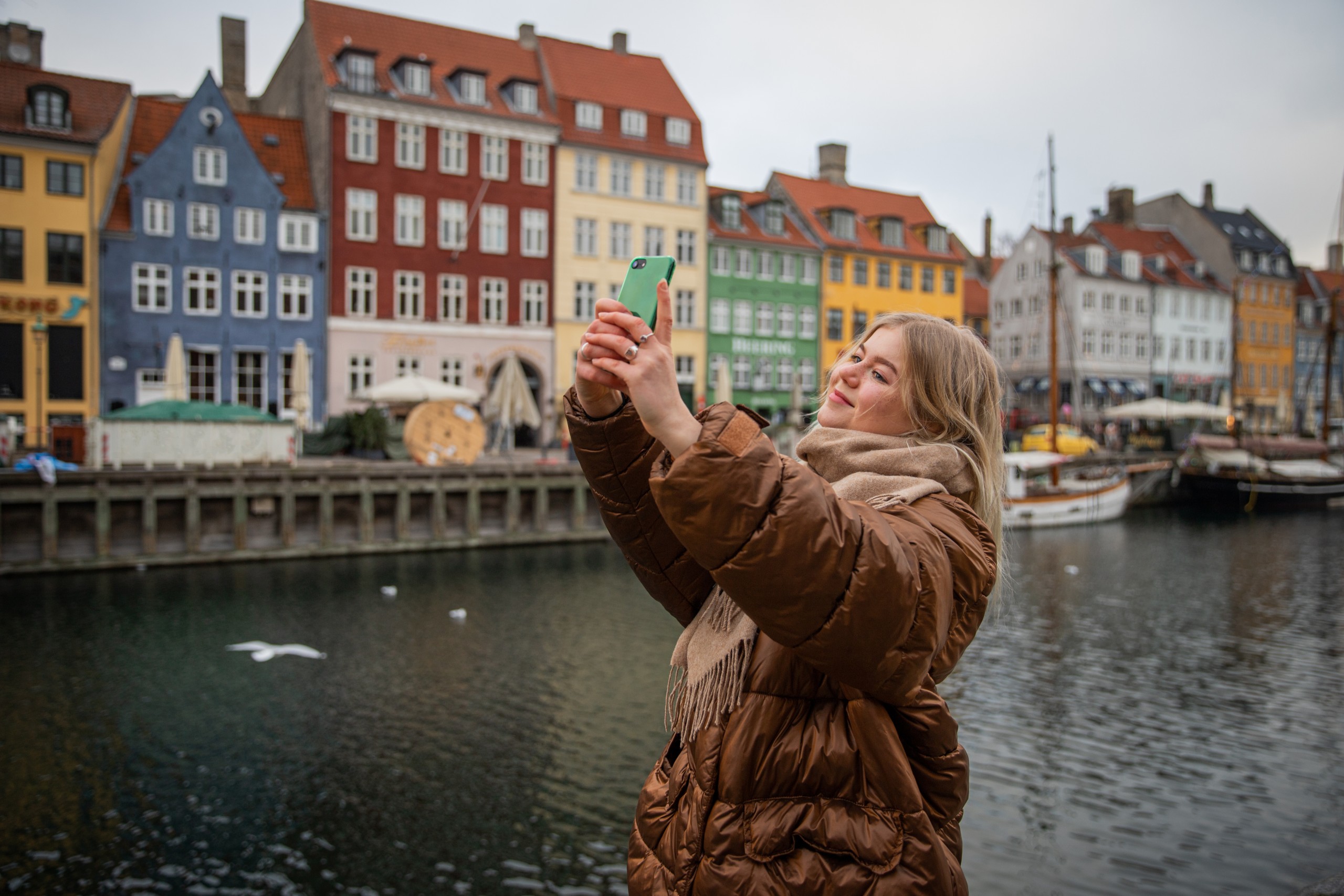 A young woman in a brown puffer jacket takes a selfie with a green-cased phone in front of the colourful canal houses of Nyhavn, Copenhagen