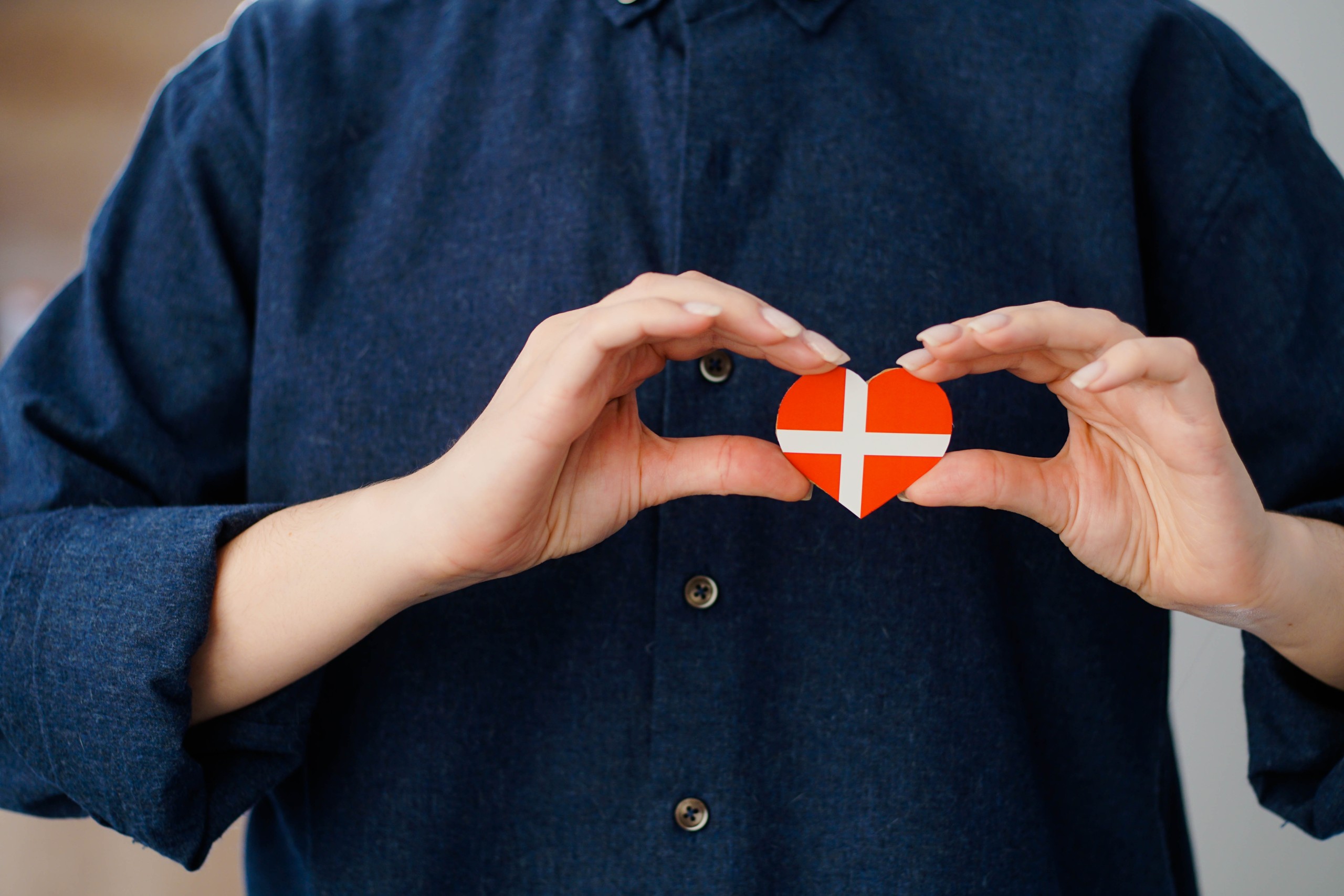 A person in a dark blue shirt holds a small heart-shaped paper cut-out printed with the Danish flag.